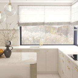 Kinga Jaźwiec - Jasna kuchnia w stylu minimalistycznym z wyspą, drewnianą misą z owocami cytrusowymi i złotą armaturą na tle okna z widokiem na łąkę.