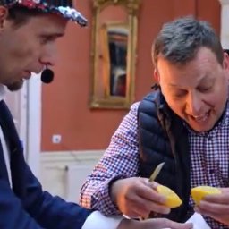 Łukasz Marczak - magik, iluzjonista - Iluzjonista w cekinowej czapce prezentuje trik z cytryną, drugi mężczyzna z zainteresowaniem obserwuje. W tle dekoracyjne wnętrze.