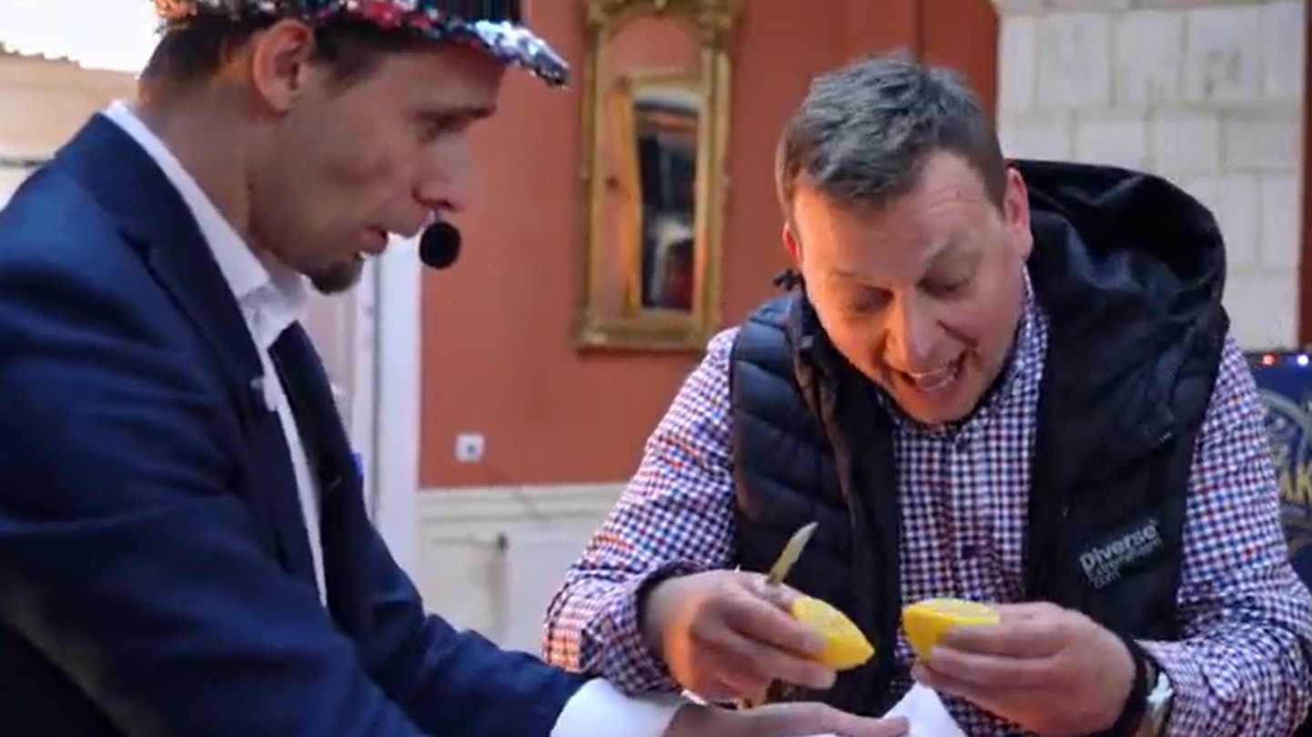 Iluzjonista w cekinowej czapce prezentuje trik z cytryną, drugi mężczyzna z zainteresowaniem obserwuje. W tle dekoracyjne wnętrze.