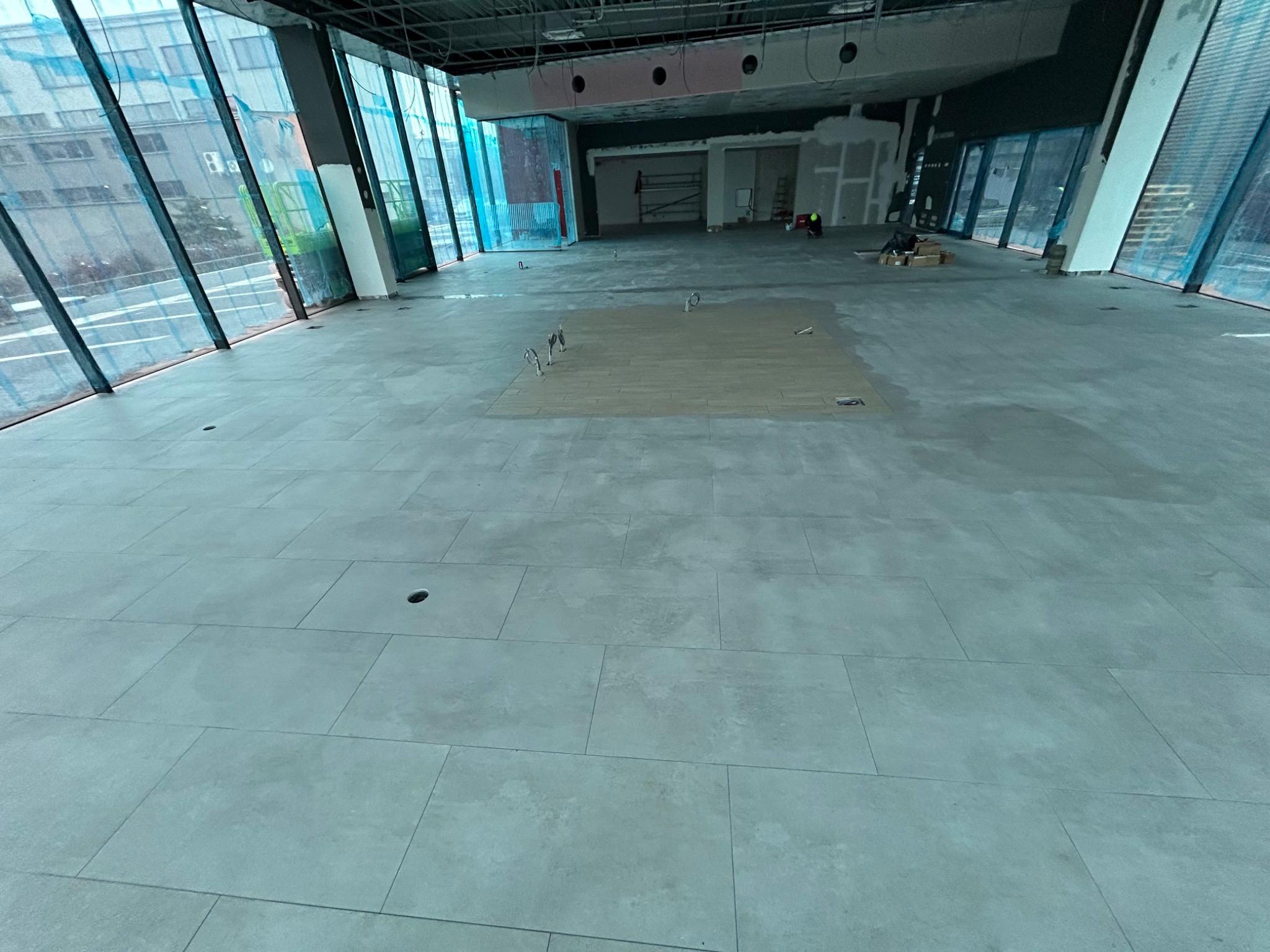 Układanie jasnoszarych płytek podłogowych w dużym, otwartym pomieszczeniu z panoramicznymi oknami; częściowo ułożony fragment z płytek imitujących drewno; widoczne narzędzia i pracownik w tle.