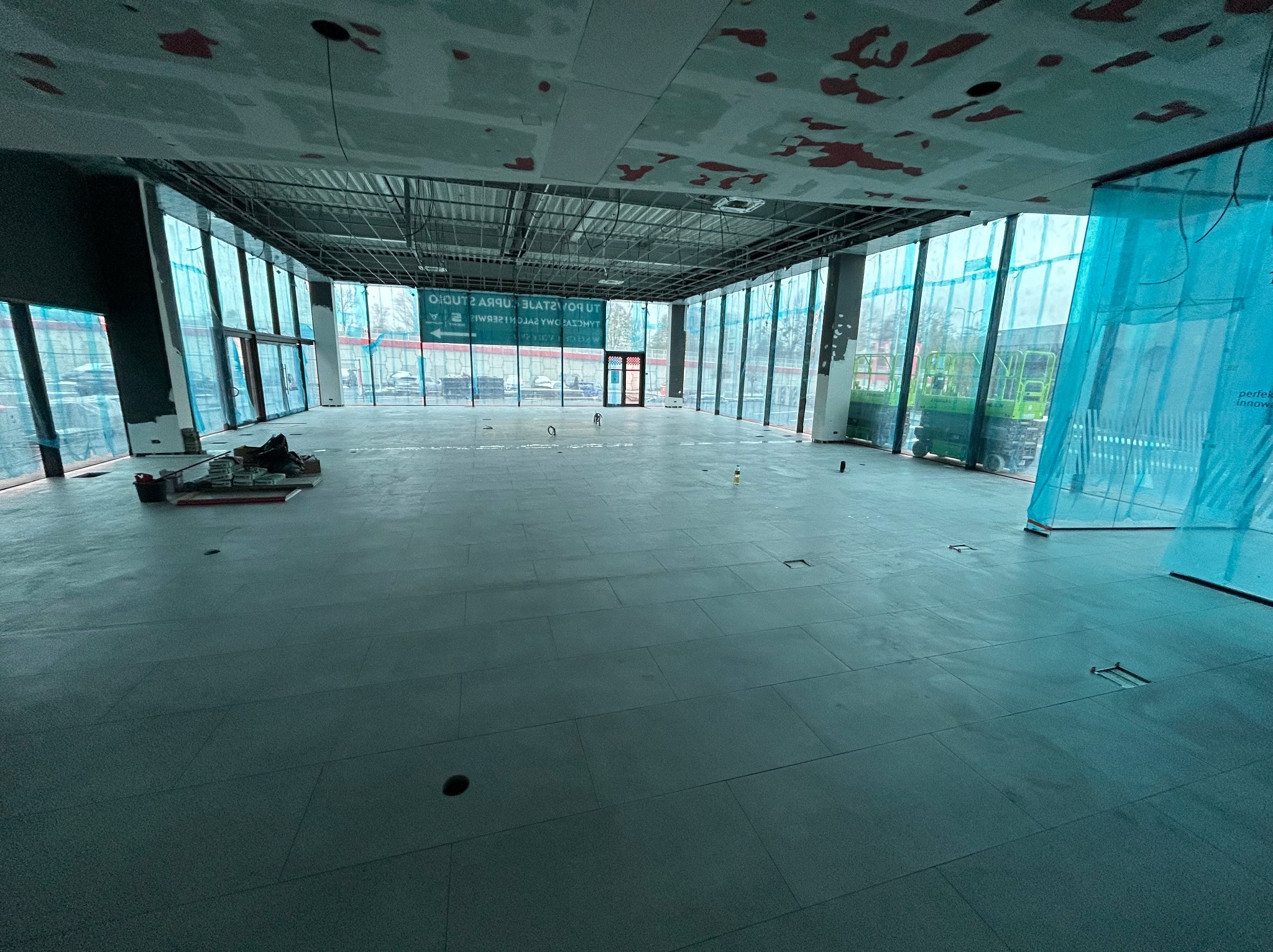 Wnętrze dużego pomieszczenia z ułożoną szarą podłogą z płytek, widoczne narzędzia pracy w rogu, ściany z dużymi oknami zabezpieczone folią, surowy sufit z instalacjami.