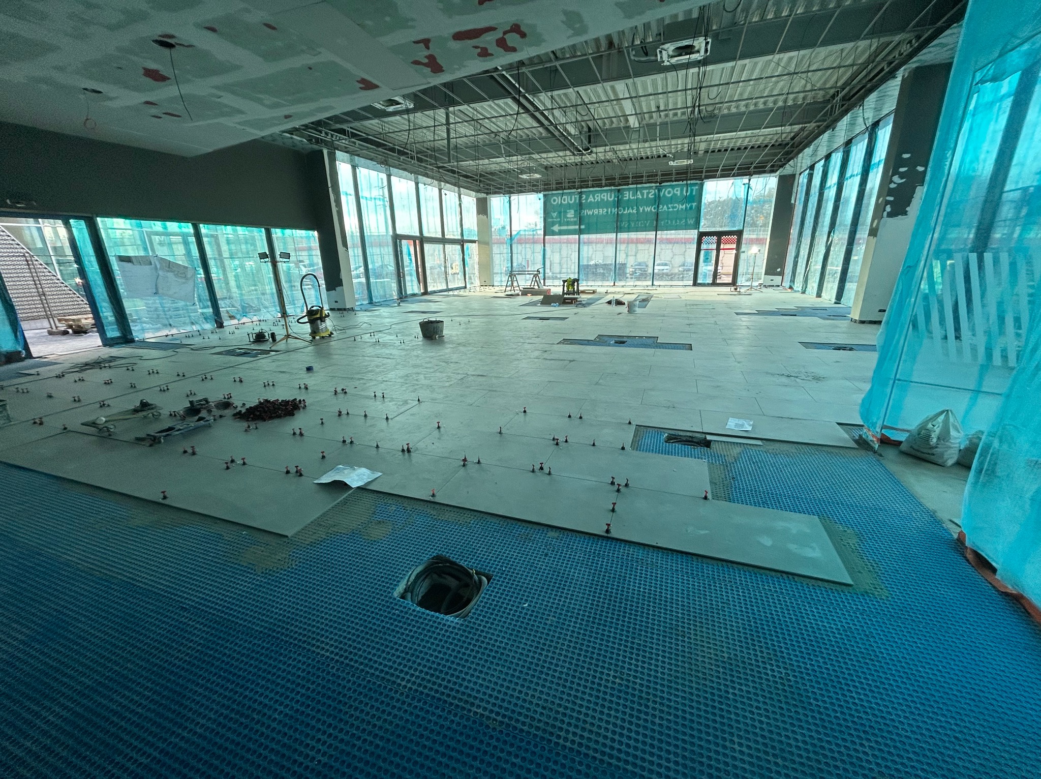 Układanie płytek podłogowych w dużym, jasnym pomieszczeniu z panoramicznymi oknami, widoczne narzędzia i system poziomowania płytek.