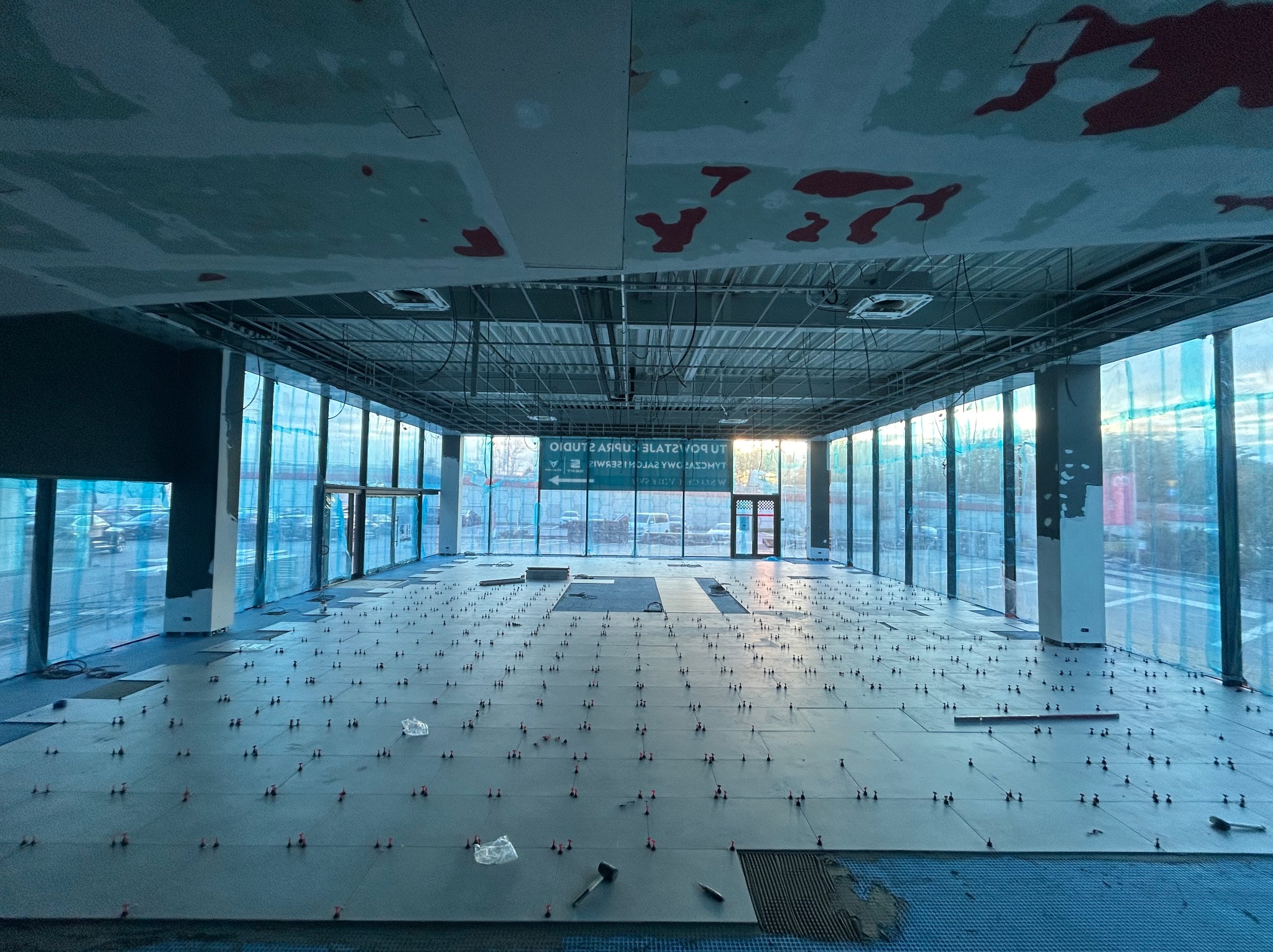 Układanie płytek podłogowych w dużym, otwartym pomieszczeniu z panoramicznymi oknami, widoczne krzyżyki dystansowe i młotek gumowy.