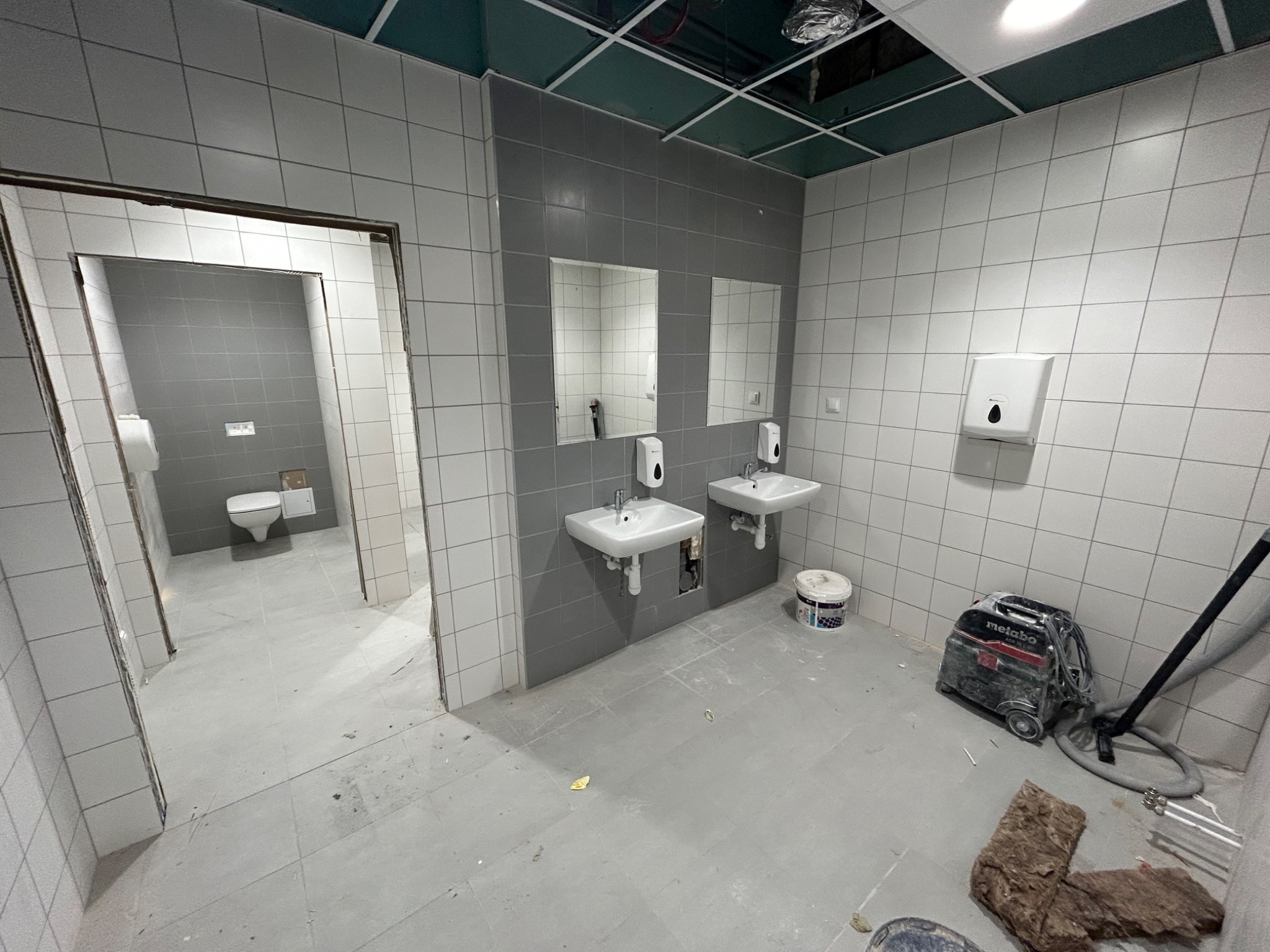 Wnętrze łazienki w trakcie remontu: szare i białe płytki, dwie umywalki z lustrami, odkurzacz przemysłowy Metabo, widoczne ślady prac wykończeniowych na podłodze.