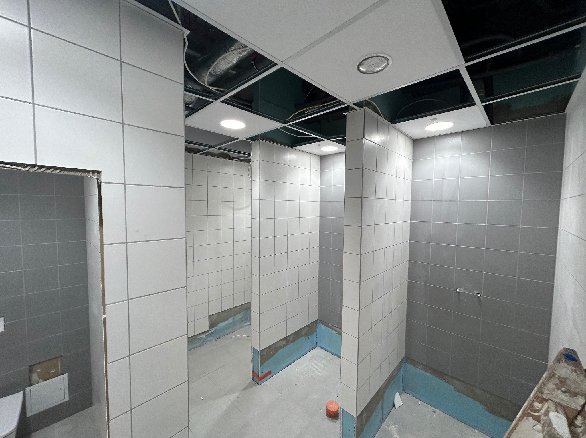 Wnętrze publicznej toalety w trakcie remontu, z częściowo ułożonymi białymi i szarymi płytkami na ścianach, widoczne zabezpieczenia hydroizolacyjne przy podłodze i sufit podwieszany z oświetleniem.