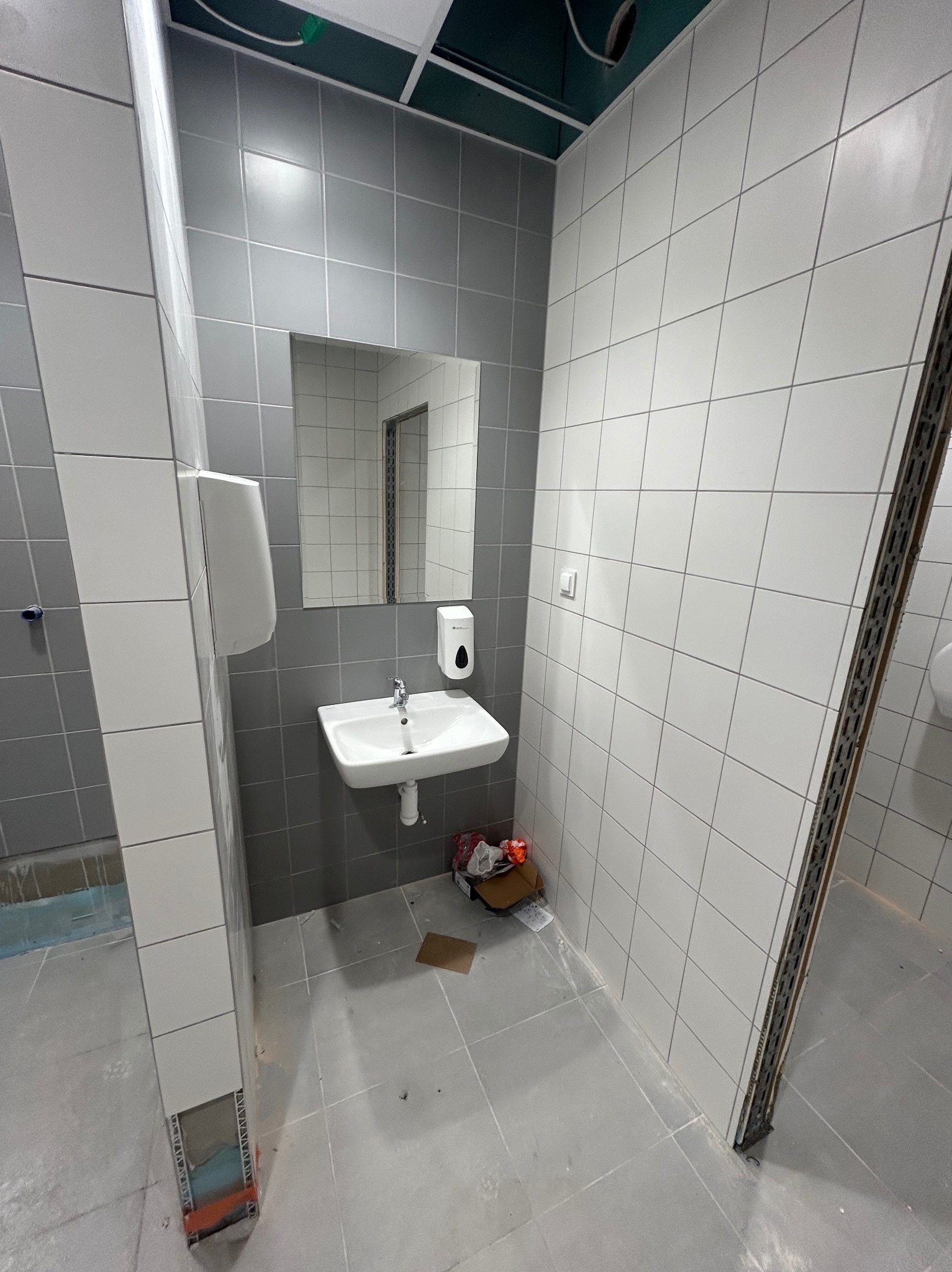 Wnętrze łazienki w trakcie remontu, widoczne szare i białe płytki na ścianach, umywalka z baterią, lustro oraz elementy montażowe na podłodze i ścianach.