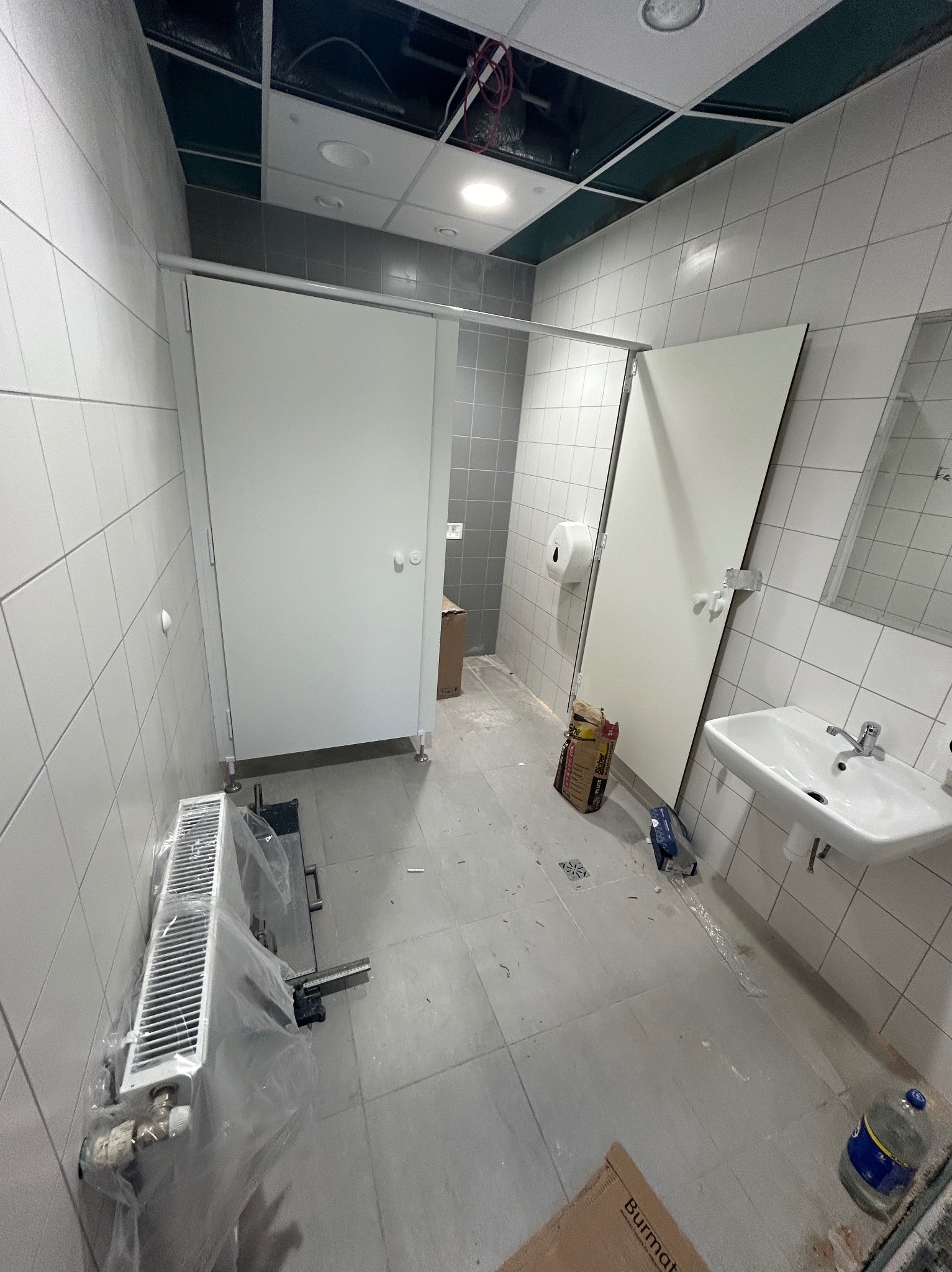 Montaż kabin sanitarnych w publicznej toalecie: widoczne białe kabiny z metalowymi okuciami, umywalka, grzejnik w folii ochronnej, płytki ceramiczne na ścianach i podłodze, sufit podwieszany...