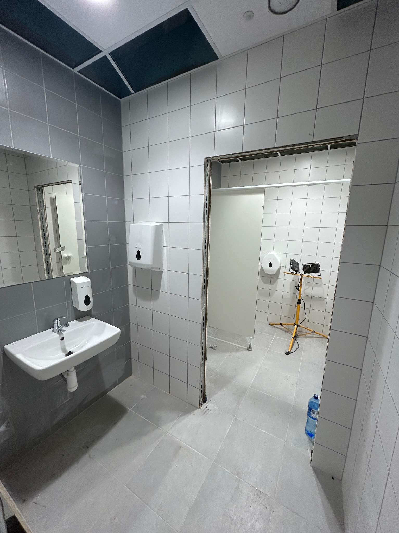 Wnętrze publicznej toalety w trakcie remontu: szare i białe płytki ceramiczne na ścianach i podłodze, umywalka z dozownikiem mydła, lustro, dozownik ręczników, tymczasowe oświetlenie na statywie...