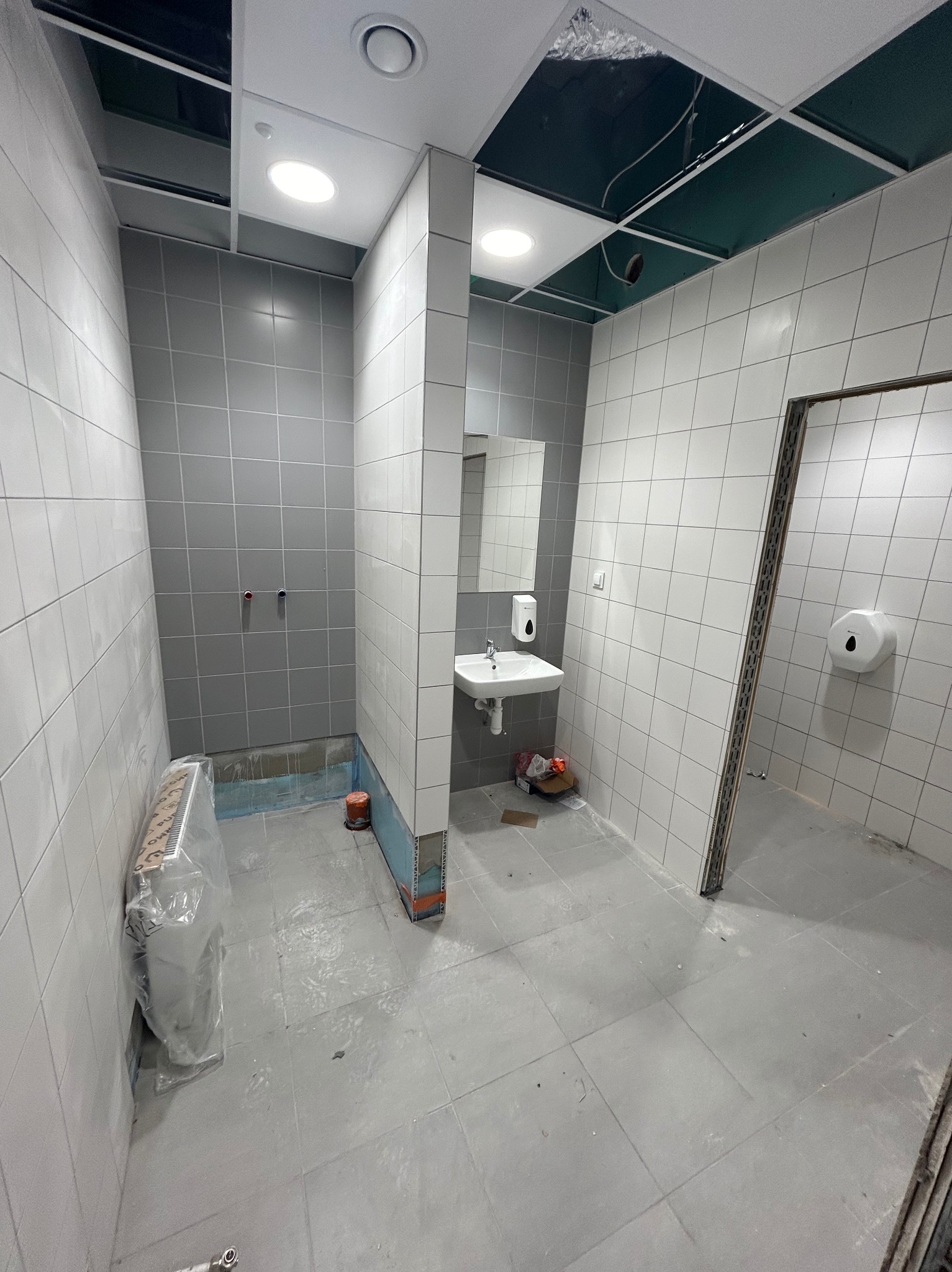 Wnętrze łazienki w trakcie remontu: szare płytki na ścianach i podłodze, umywalka z dozownikiem mydła, grzejnik owinięty folią, widoczne niedokończone sufity podwieszane.