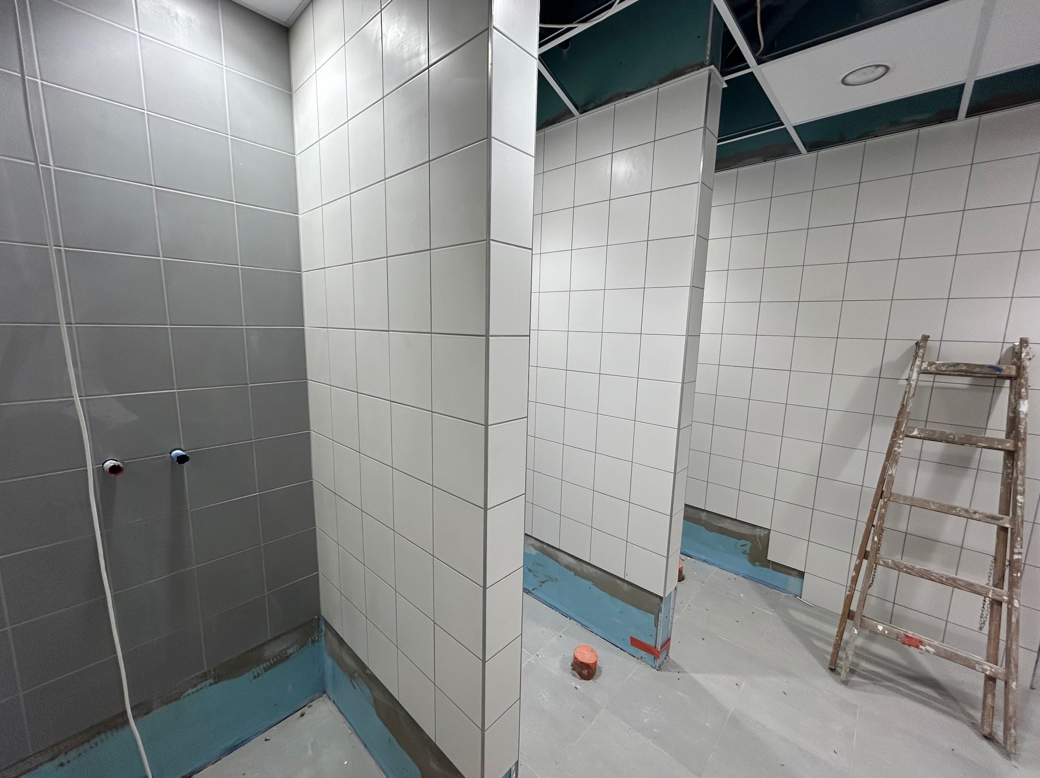 Wnętrze łazienki w trakcie układania białych płytek kwadratowych na ścianach, widoczne niebieskie zabezpieczenie przeciwwilgociowe przy podłodze, drabina malarska i wystające rury instalacji wodnej.