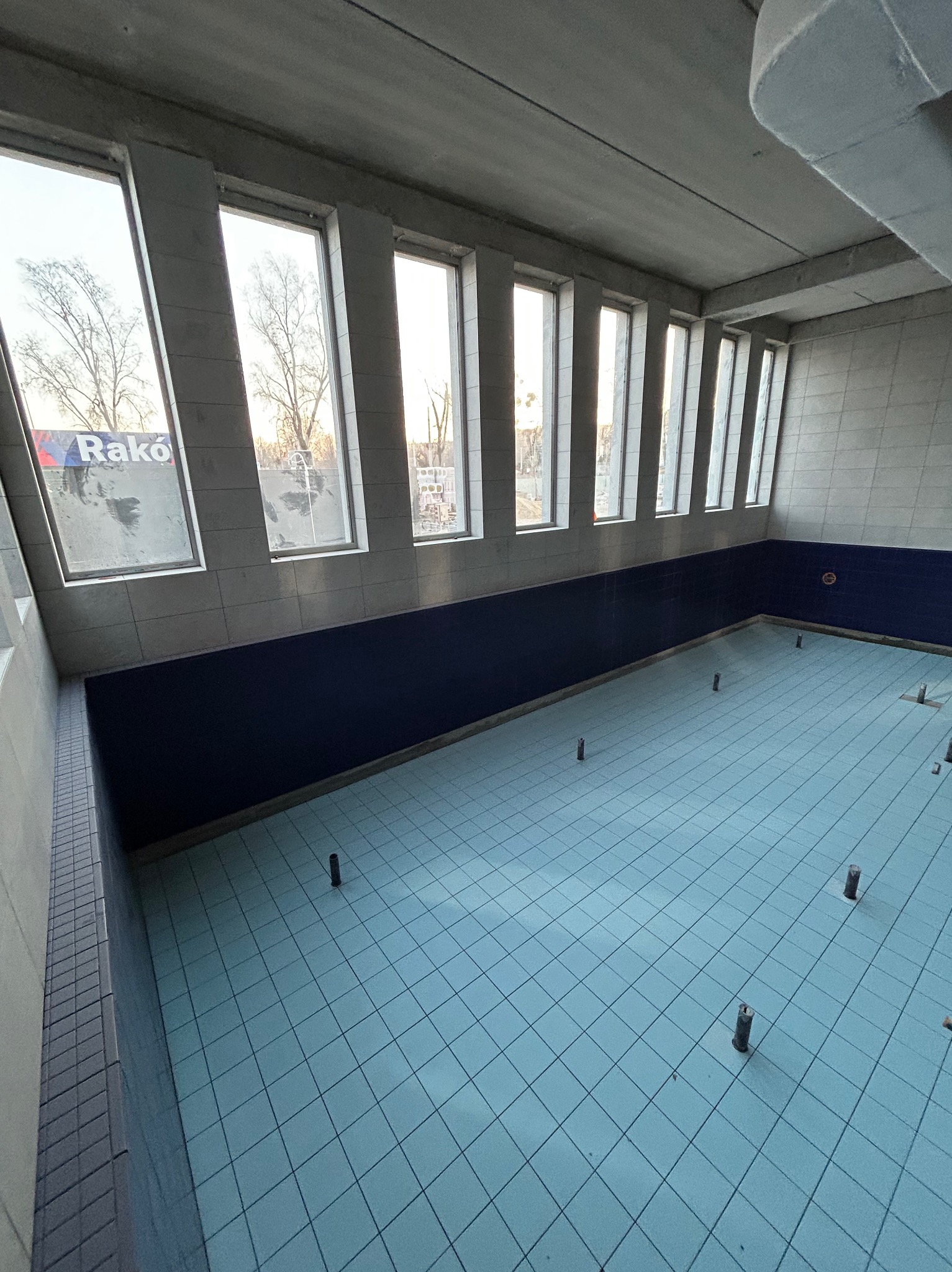 Pusty basen w trakcie remontu z niebieską glazurą i wystającymi elementami instalacji, widok z góry w kierunku okien z reklamą Rako.