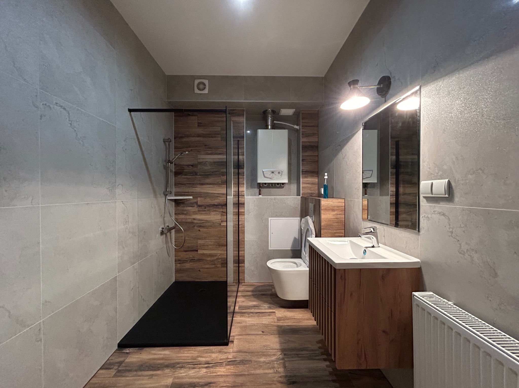 Wykończona łazienka z kabiną prysznicową typu walk-in z czarnym brodzikiem, umywalką z szafką z lamelami, podwieszaną toaletą i grzejnikiem panelowym, ściany wyłożone szarymi płytkami imitującymi...