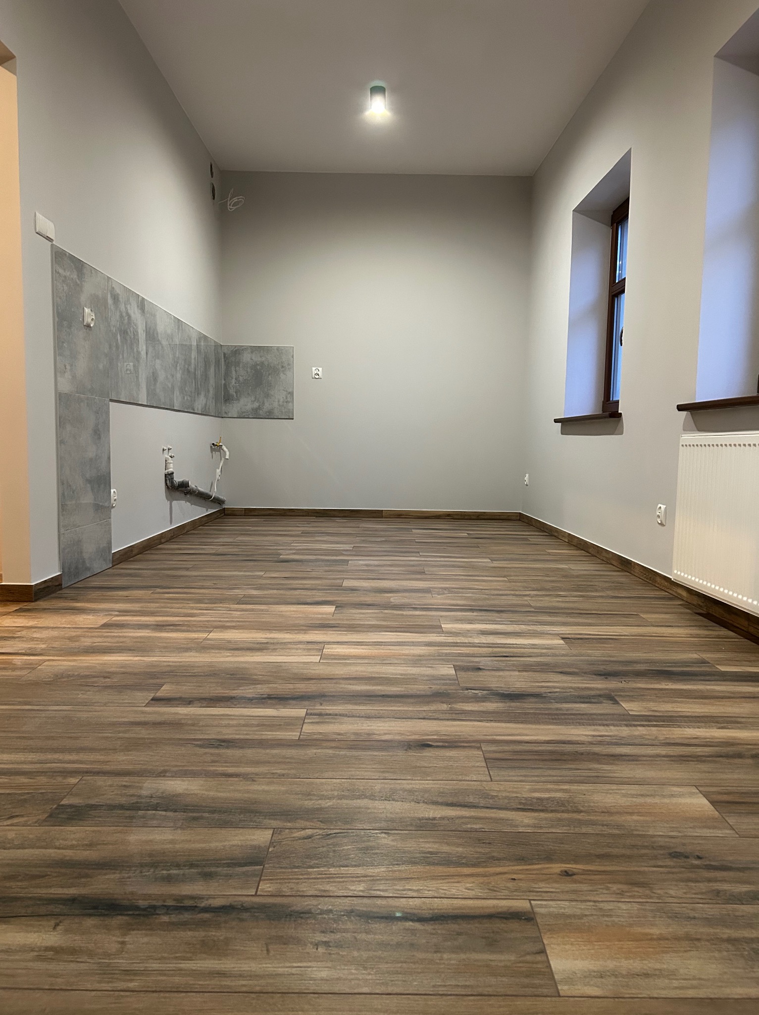 Wnętrze pomieszczenia w trakcie remontu: częściowo położone szare płytki na ścianie, drewnopodobna podłoga z płytek, grzejnik pod oknem, widoczne instalacje wodno-kanalizacyjne.