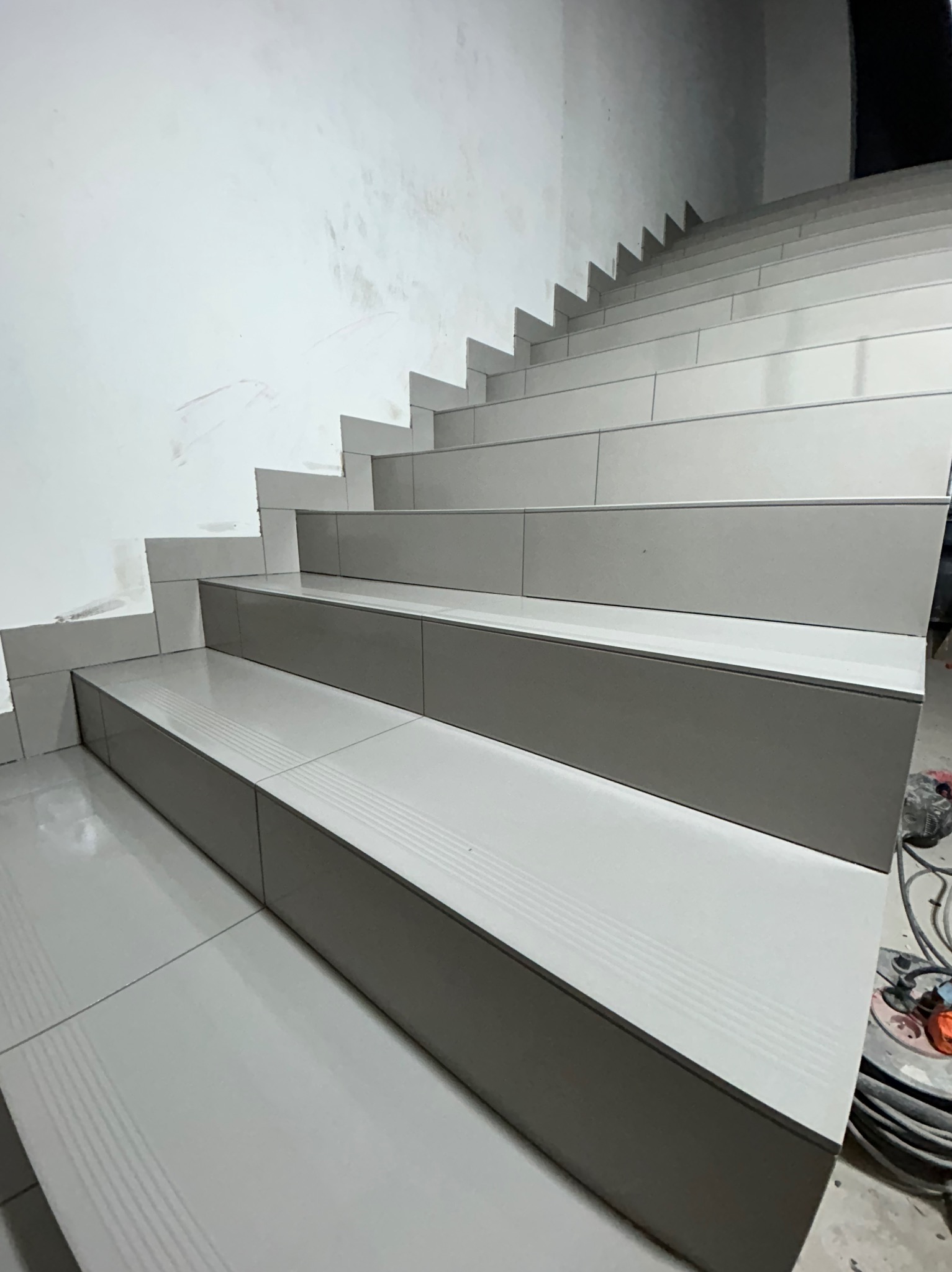 Schody wyłożone błyszczącymi, szarymi płytkami z delikatnym wzorem, widoczne ślady po pracach wykończeniowych na ścianie i narzędzia w tle.