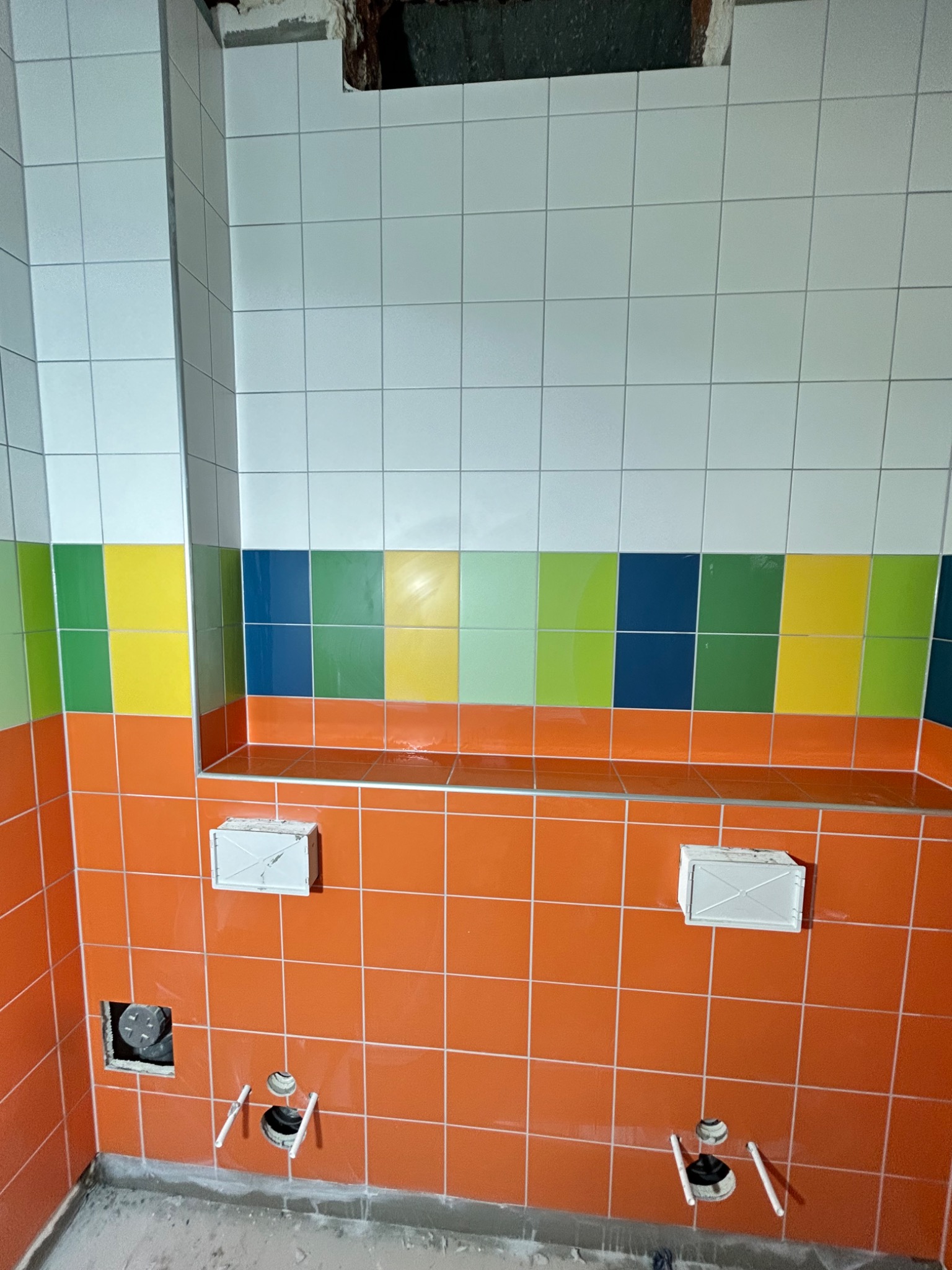 Ściana w łazience w trakcie wykańczania, z pomarańczowymi płytkami na dole, pasem kolorowych płytek (zielony, żółty, niebieski) i białymi płytkami u góry, widoczne otwory na instalacje, szare fugi.