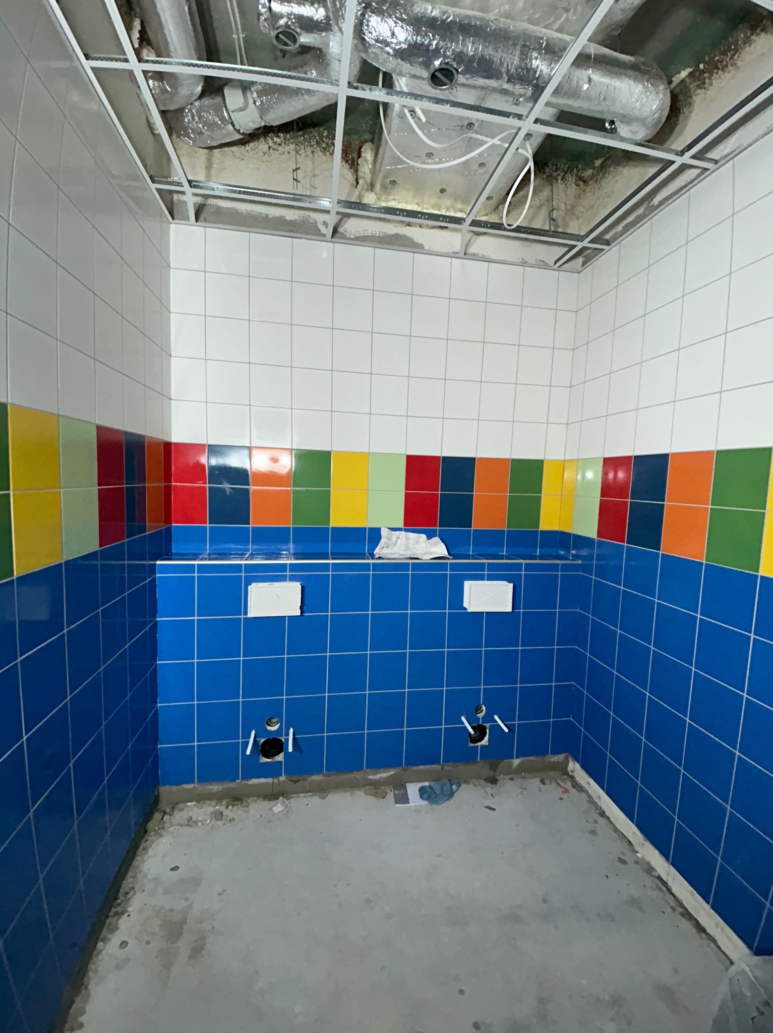 Niebieskie płytki w łazience z kolorowym pasem na ścianie, surowy beton na podłodze i niezabudowany sufit z widoczną wentylacją.