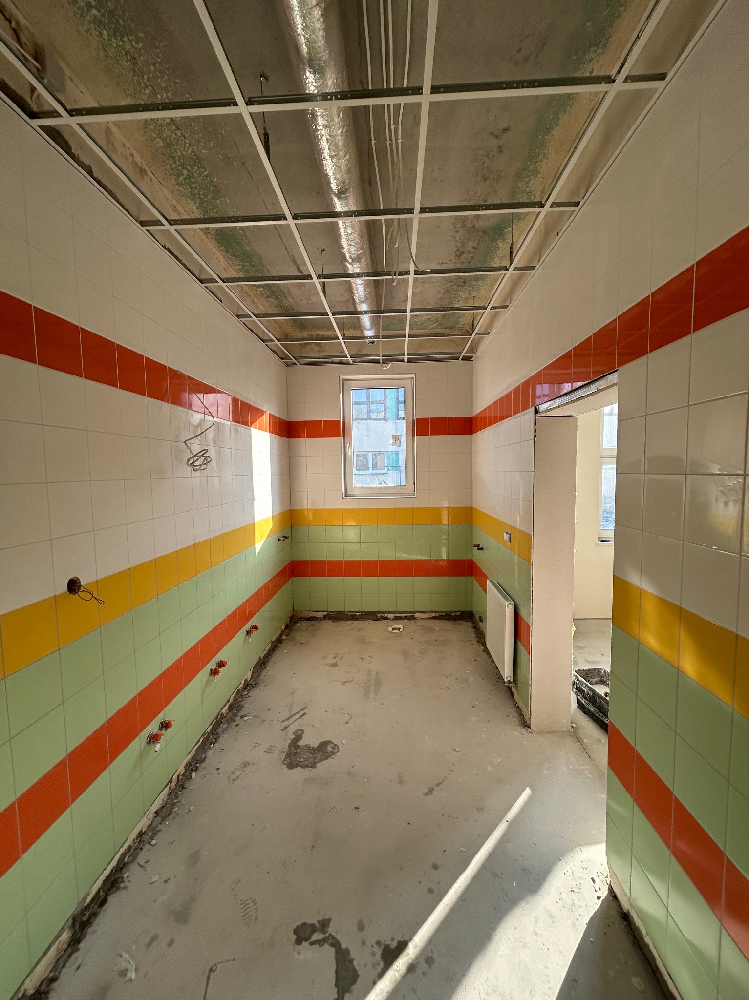 Wnętrze w trakcie remontu z kolorowymi pasami płytek (pomarańczowy, żółty, zielony) na ścianach, sufit w trakcie montażu, widoczne instalacje elektryczne i hydrauliczne, surowa podłoga.