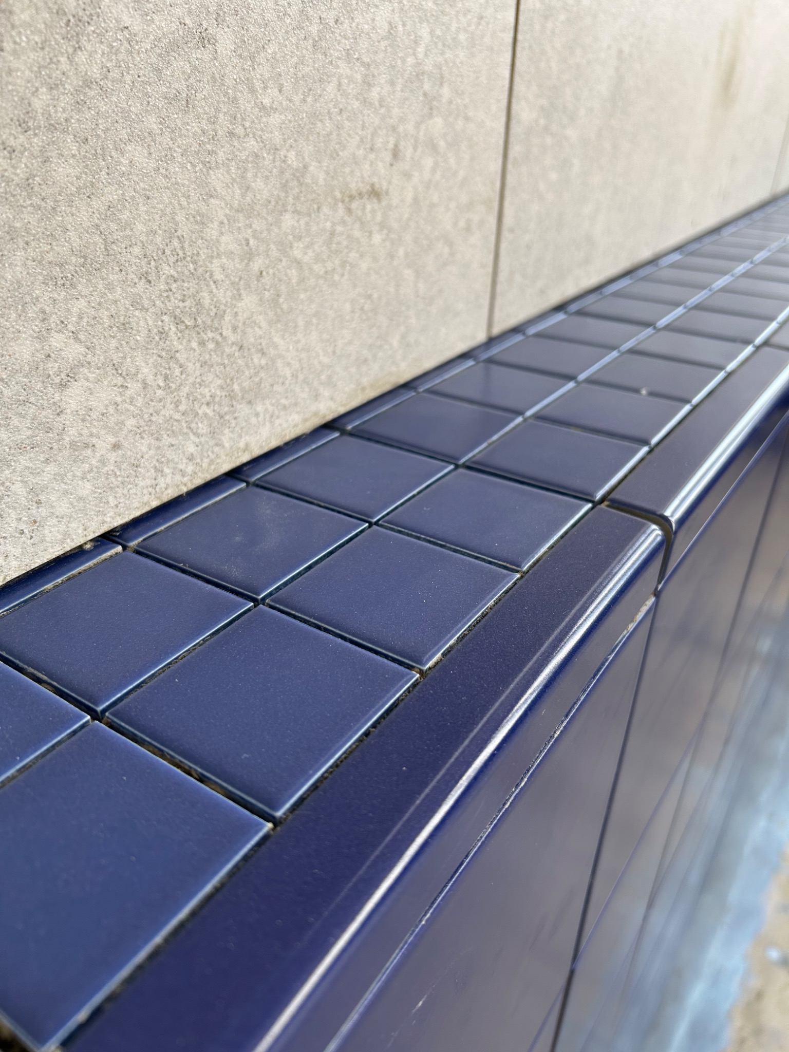 Niebieskie, błyszczące płytki ceramiczne ułożone na krawędzi ściany z szarego betonu, widoczne spoiny między płytkami.