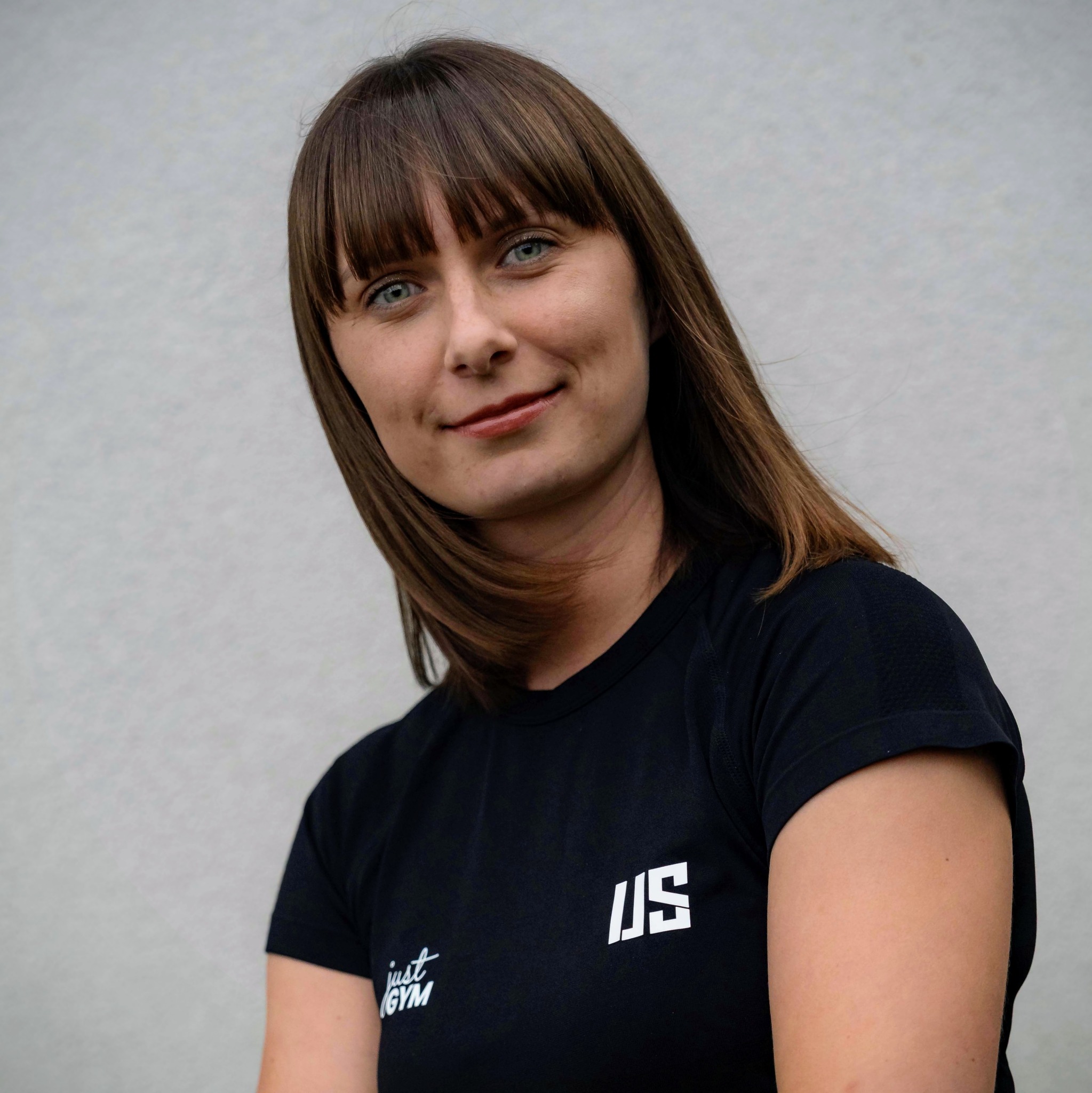 Portret uśmiechniętej kobiety z niebieskimi oczami w czarnej koszulce Just Gym na tle jasnej ściany. Zdjęcie promujące siłownię.