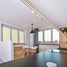 Quality Apartments - Nowoczesna kuchnia z czarnym blatem, zlewozmywakiem i baterią, jasne meble i stół z krzesłami w tle, oświetlenie szynowe i okap podszafkowy.