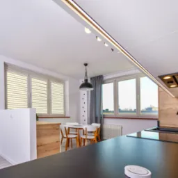 Nowoczesna kuchnia z czarnym blatem, zlewozmywakiem i baterią, jasne meble i stół z krzesłami w tle, oświetlenie szynowe i okap podszafkowy.