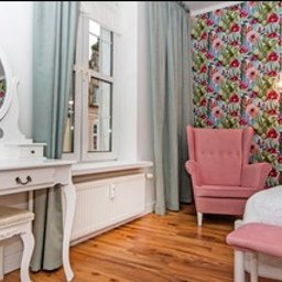 Quality Apartments - Jasny pokój z białym biurkiem z lustrem, różowym fotelem na tle ściany w kwiaty i drewnianą podłogą.