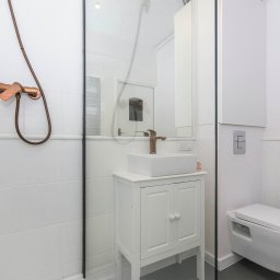 Quality Apartments - Nowoczesna łazienka z białą ceramiką, kabiną prysznicową ze szklaną ścianką, umywalką na szafce i armaturą w kolorze miedzi.