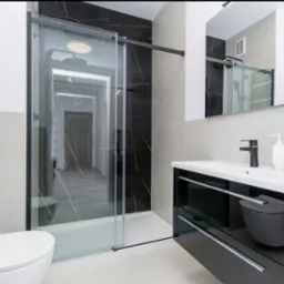 Nowoczesna łazienka z czarnymi marmurowymi akcentami, kabiną prysznicową typu walk-in z przesuwnymi drzwiami, umywalką nablatową na czarnej szafce z chromowanymi uchwytami i wiszącą toaletą.