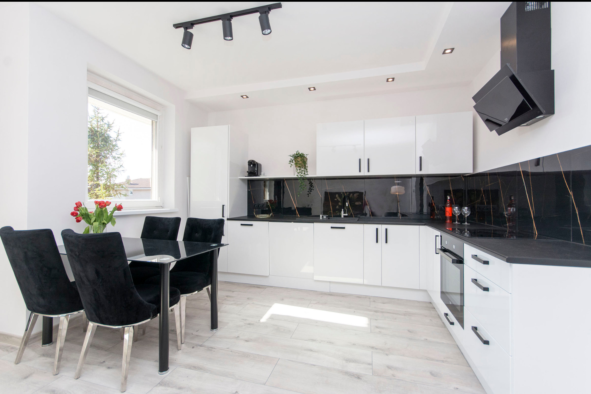 Nowoczesna, jasna kuchnia z białymi szafkami, czarnym blatem i marmurowym backsplashiem, czarny stół z welurowymi krzesłami i bukietem czerwonych tulipanów.
