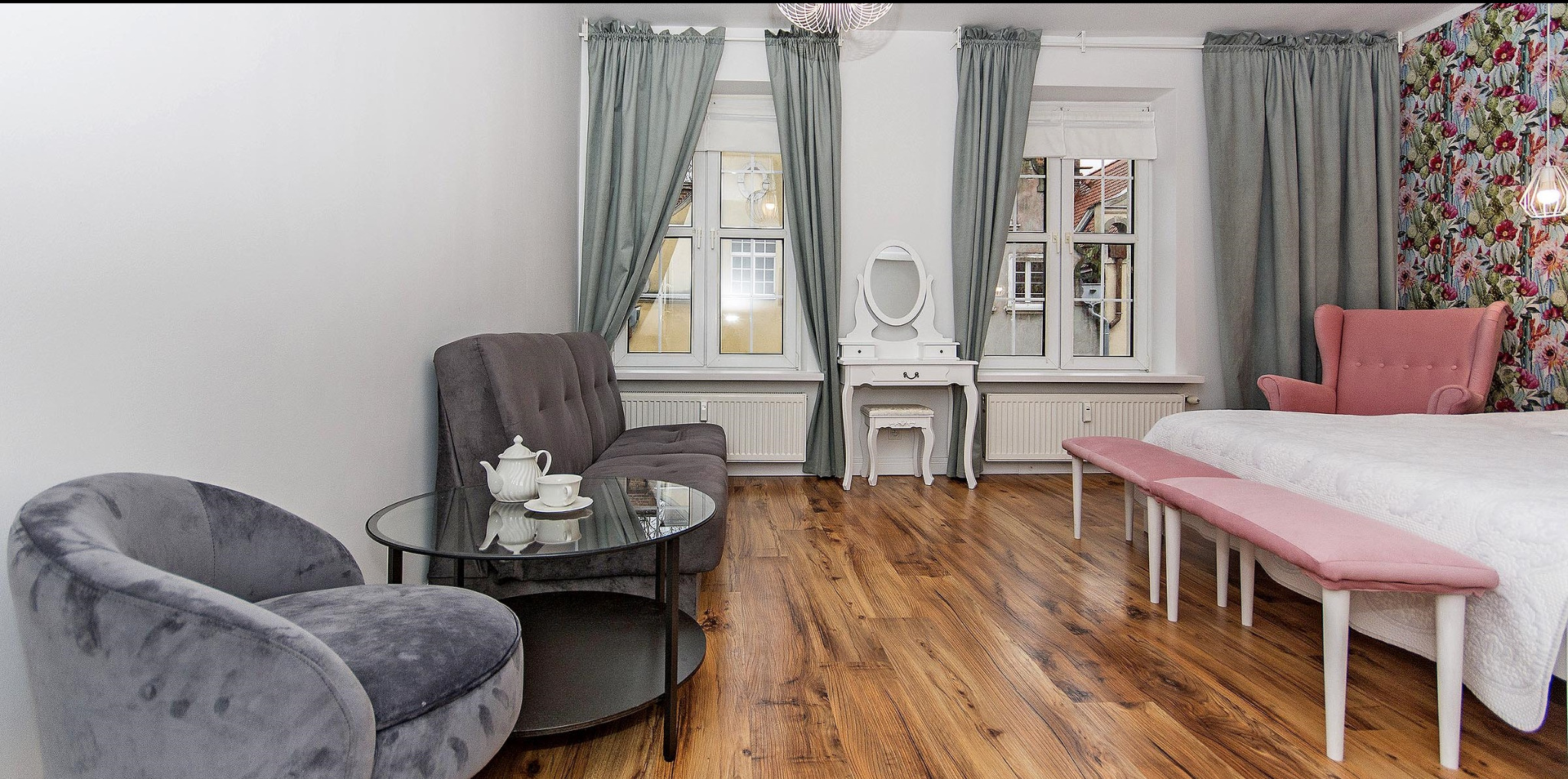 Pokój z dwoma oknami z szarymi zasłonami, szarym fotelem i sofą, okrągłym szklanym stolikiem z porcelanowym czajnikiem i filiżanką, białą toaletką z lustrem, różowym fotelem i ławką oraz wzorzystą...
