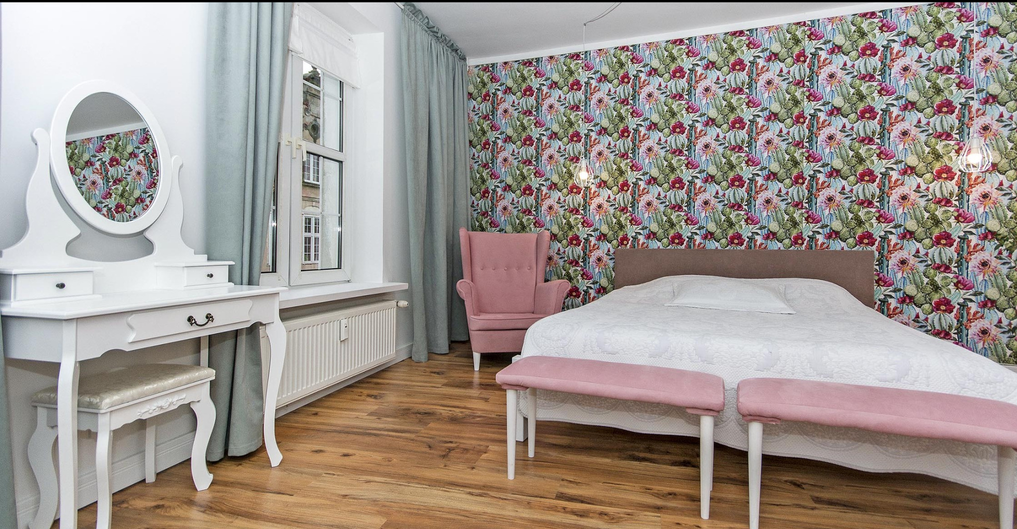 Pokój z łóżkiem, toaletką z owalnym lustrem, różowym fotelem i tapetą w kaktusy i kwiaty; podwieszana lampa z żarówką Edisona.