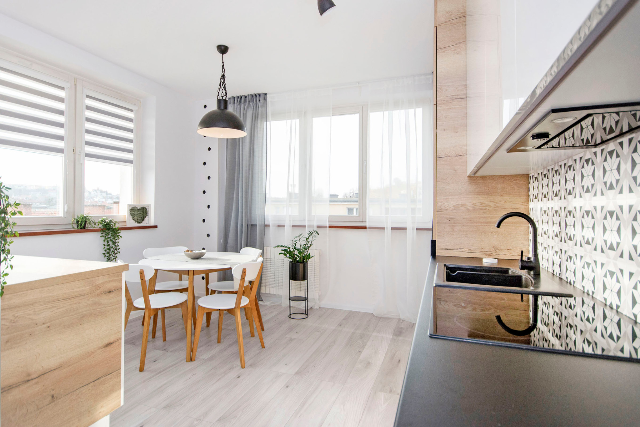 Jasna kuchnia z okrągłym stołem, białymi krzesłami i czarnym zlewem na tle wzorzystych płytek.