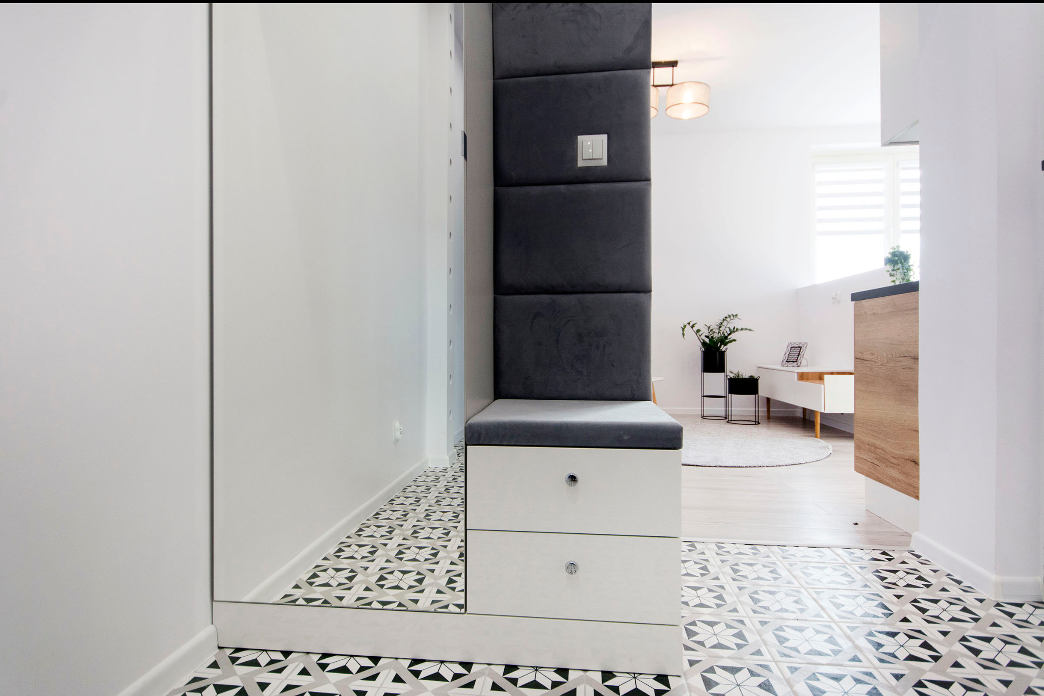 Nowoczesny przedpokój z lustrem, siedziskiem z szufladami i tapicerowanym panelem ściennym w kolorze grafitowym, wzorzysta podłoga w odcieniach szarości i bieli.