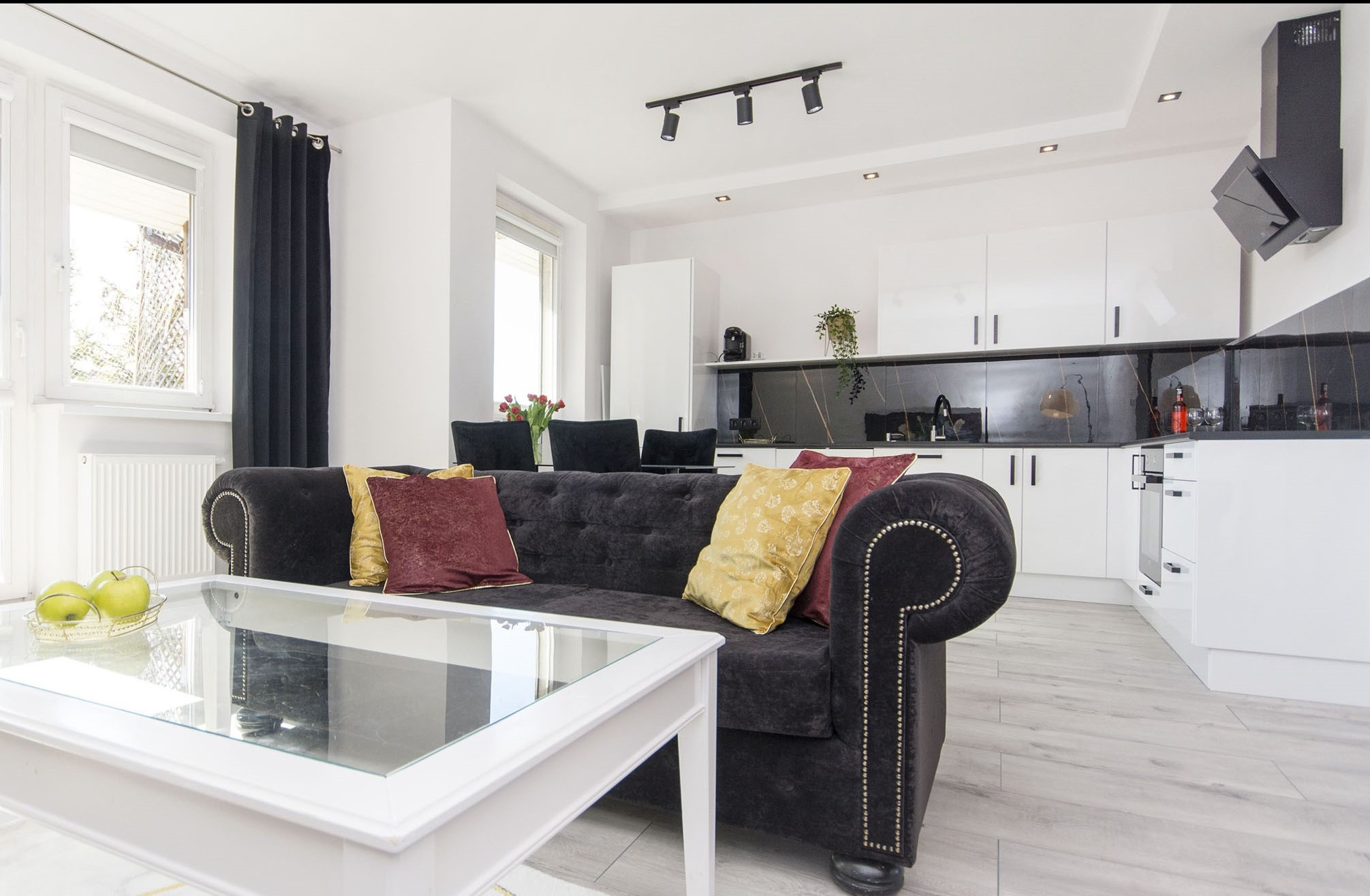 Nowoczesne wnętrze: czarna sofa Chesterfield z poduszkami w odcieniach złota i bordo na tle białej kuchni z czarnym blatem, szklany stolik kawowy z zielonymi jabłkami, czarne zasłony w oknie.