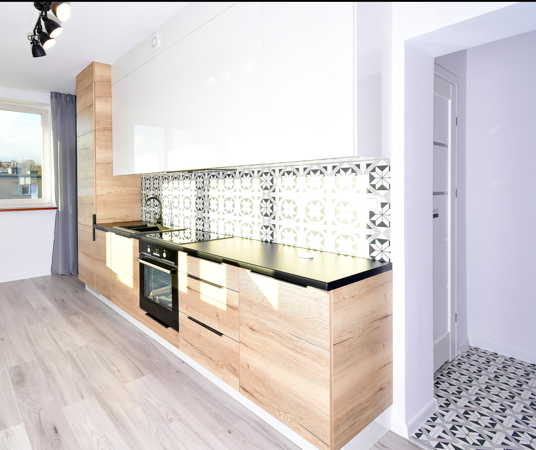 Nowoczesna kuchnia z drewnianymi szafkami dolnymi, białymi górnymi, czarnym blatem i wzorzystymi płytkami nad blatem, widok na korytarz z identycznymi płytkami na podłodze.