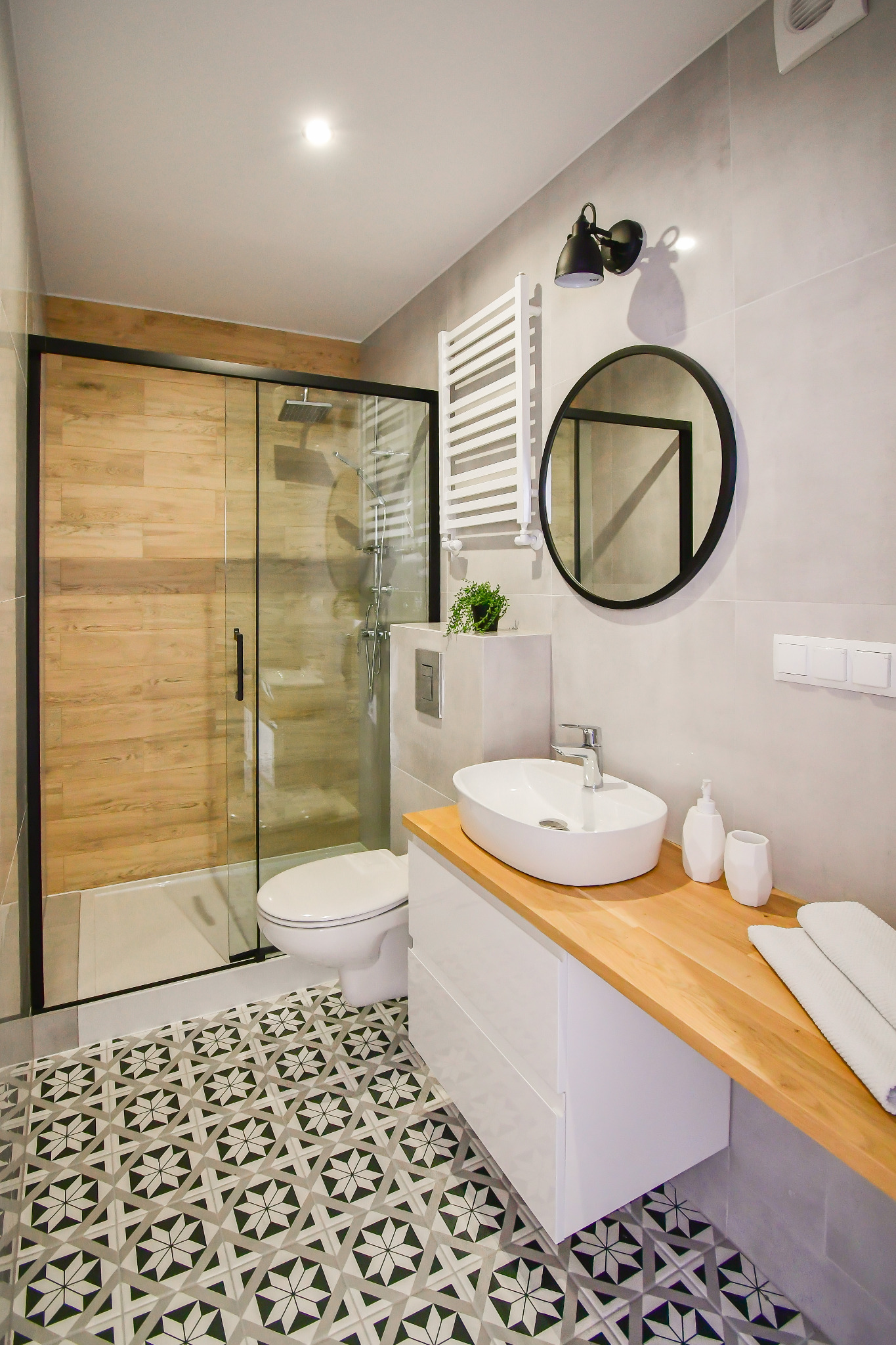 Nowoczesna łazienka z kabiną prysznicową z drewnopodobnymi panelami, umywalką nablatową na drewnianym blacie, okrągłym lustrem i wzorzystą podłogą.