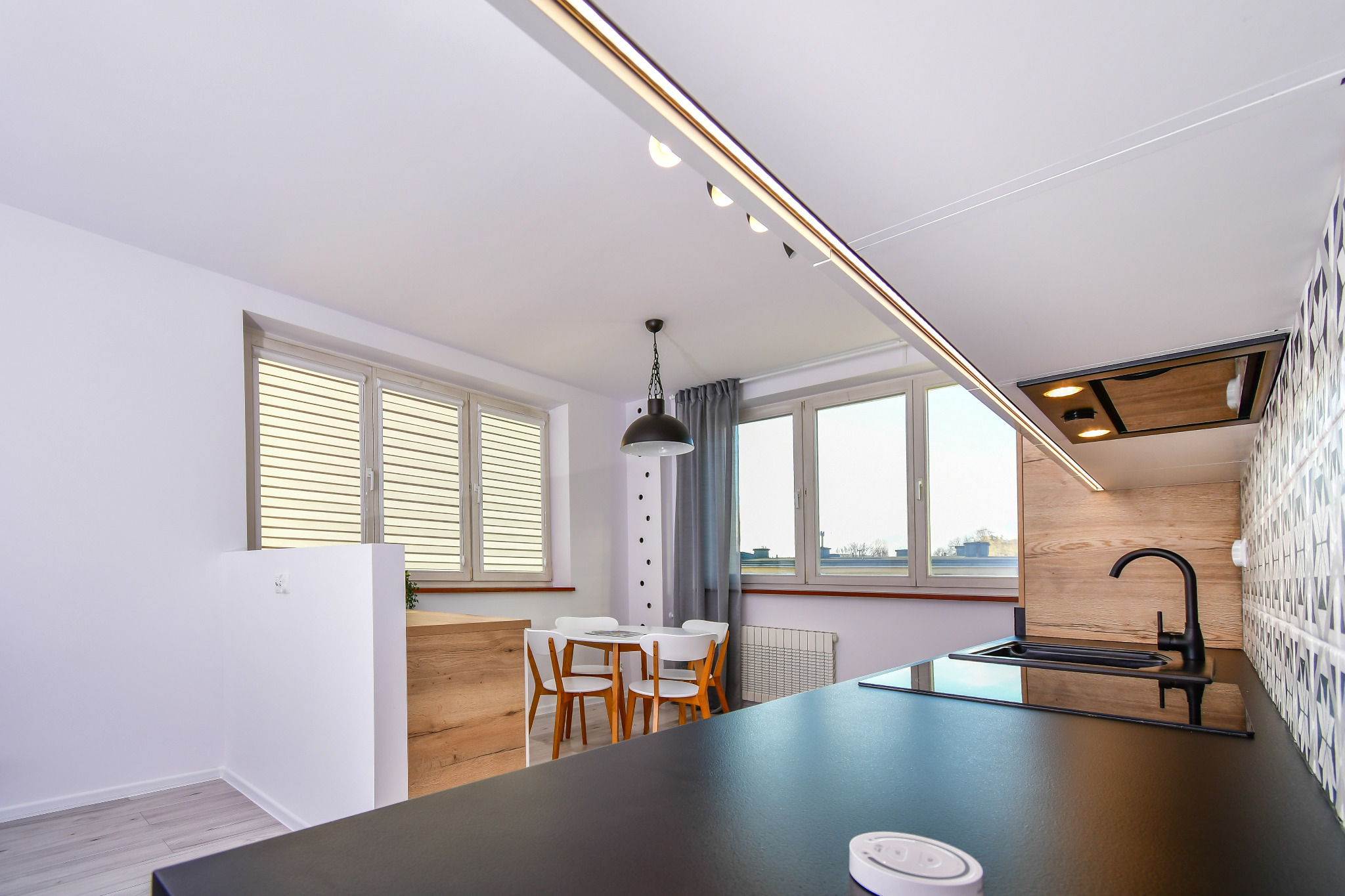 Nowoczesna kuchnia z czarnym blatem, zlewozmywakiem i baterią, jasne meble i stół z krzesłami w tle, oświetlenie szynowe i okap podszafkowy.