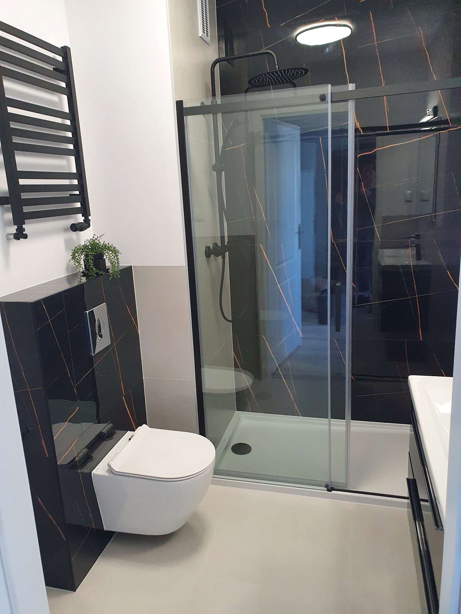 Nowoczesna łazienka z czarnymi płytkami imitującymi marmur, kabiną prysznicową typu walk-in z czarnymi elementami, podwieszaną toaletą i czarnym grzejnikiem drabinkowym.