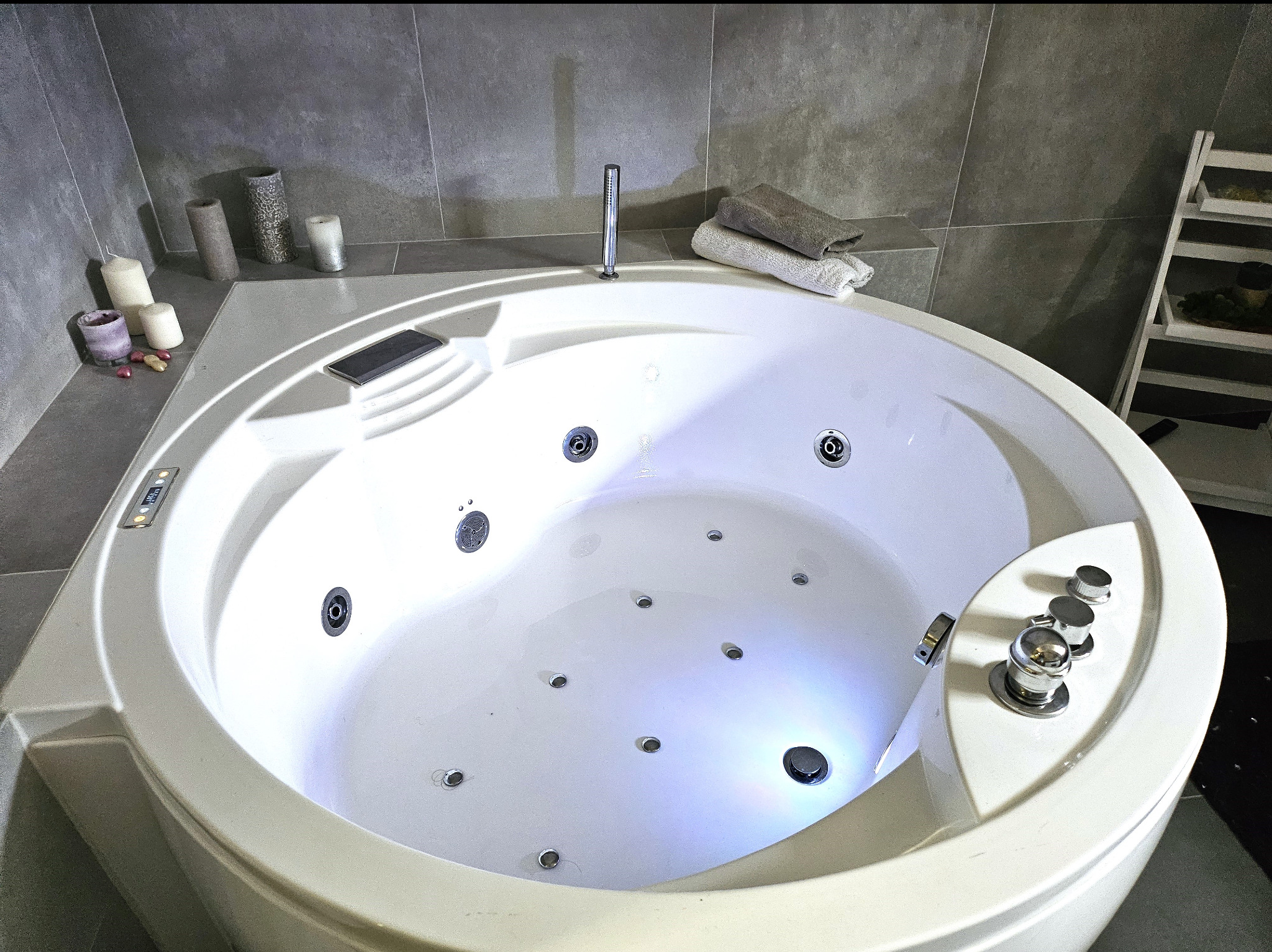 Okrągła, biała wanna z hydromasażem i podświetleniem LED, ustawiona w łazience wykończonej szarą płytką, z dekoracyjnymi świecami i ręcznikami w tle.