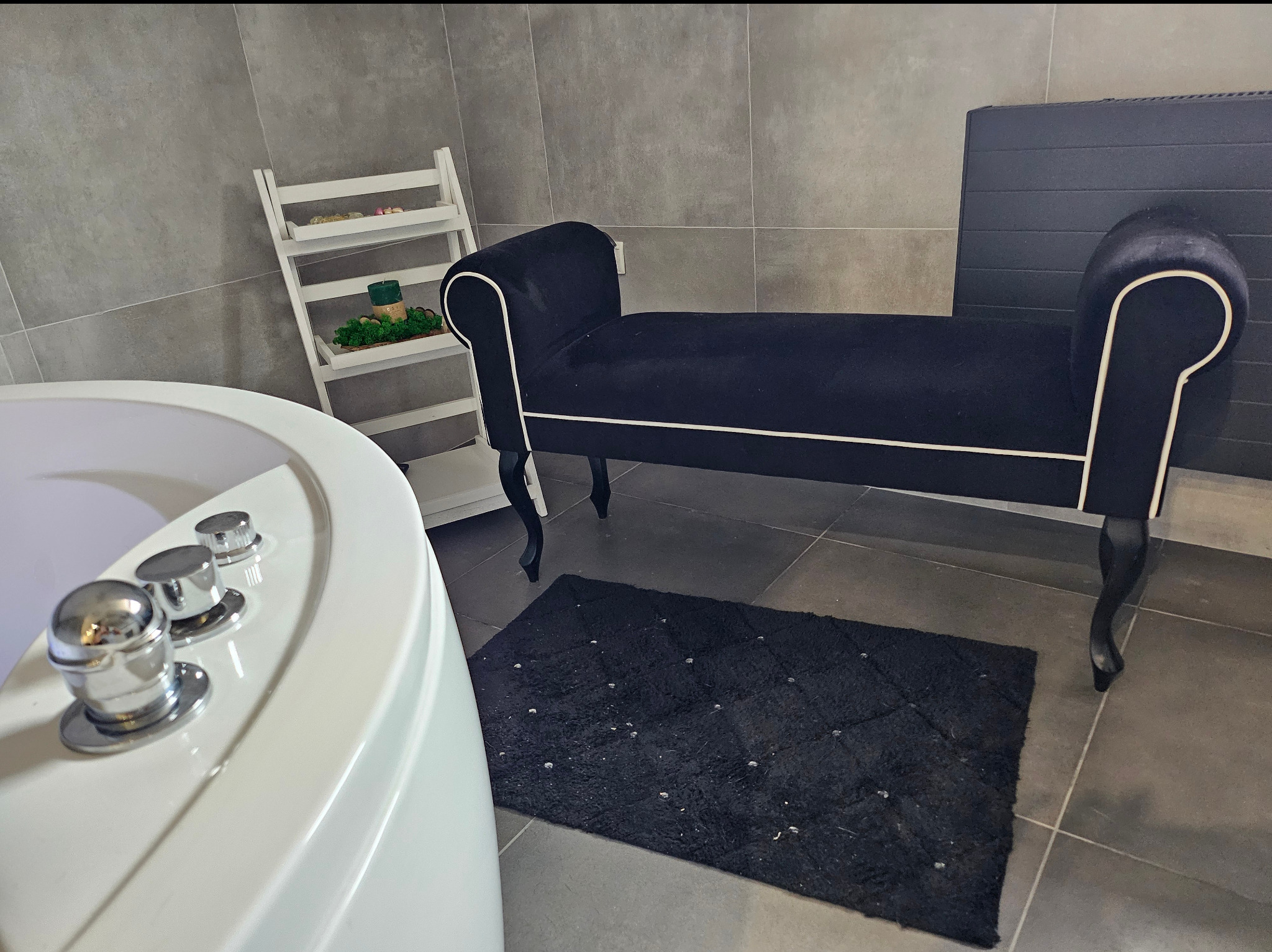 Nowoczesna łazienka z wolnostojącą wanną z hydromasażem, czarną ławką tapicerowaną białym sznurkiem i czarnym dywanikiem z długim włosiem.