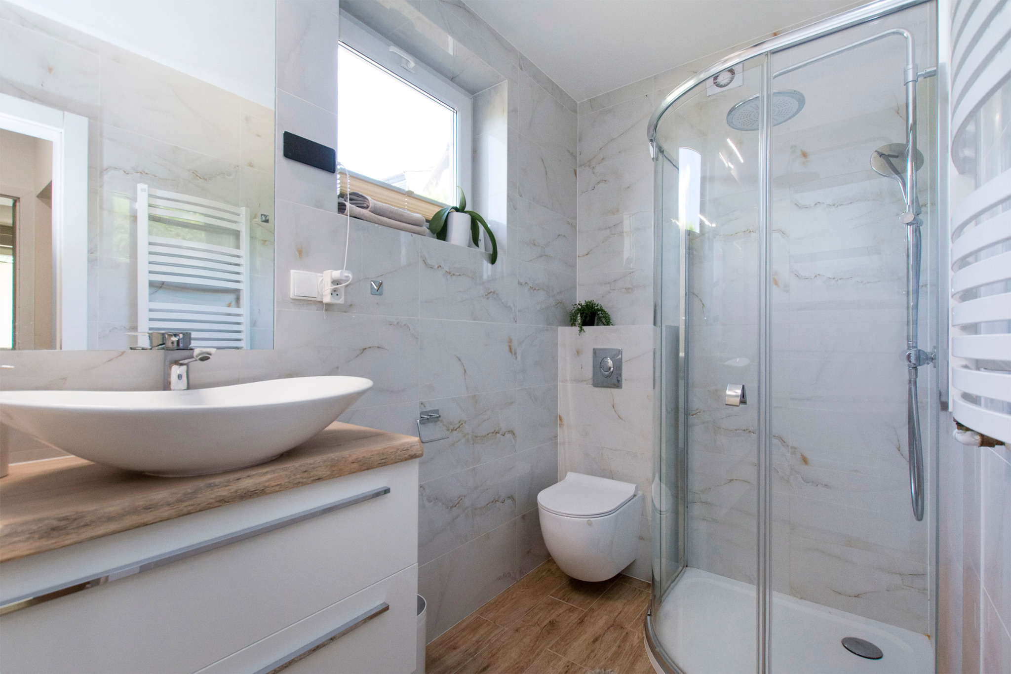 Jasna łazienka z marmurowymi płytkami, owalną umywalką na drewnianym blacie, kabiną prysznicową i wiszącą toaletą.