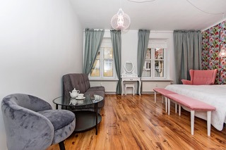 Jasne wnętrze pokoju z drewnianą podłogą, szarą sofą, okrągłym stolikiem kawowym z serwisem do herbaty, toaletką z lustrem przy oknie oraz łóżkiem z różowym wezgłowiem i tapicerowaną ławką.