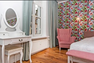 Jasny pokój z białym biurkiem z lustrem, różowym fotelem na tle ściany w kwiaty i drewnianą podłogą.
