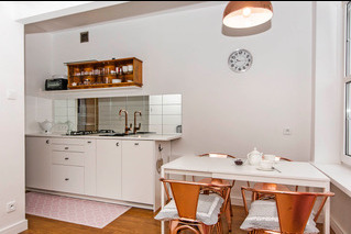 Jasna kuchnia z białymi szafkami, drewnianą półką na przyprawy, białym stołem i miedzianymi krzesłami. Nad stołem wisi miedziana lampa, a na ścianie zegar.