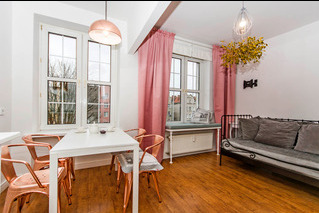 Jasne wnętrze pokoju dziennego z drewnianą podłogą, białymi ścianami i oknami. Biały stół z miedzianymi krzesłami. W tle sofa z szarymi poduszkami i dekoracje okienne z różowymi zasłonami.