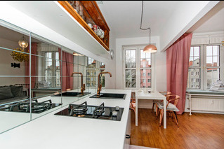 Nowoczesna kuchnia z białym blatem i lustrzanym panelem na ścianie, widoczne czarne płyty grzewcze i miedziane baterie, w tle stół z krzesłami i okno z różowymi zasłonami.