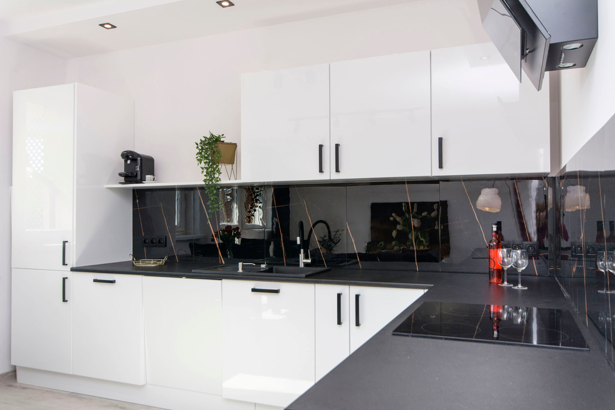 Nowoczesna kuchnia z białymi lakierowanymi szafkami, czarnym blatem z marmurowym wzorem, czarną armaturą i osprzętem, z butelką wina i kieliszkami na blacie
