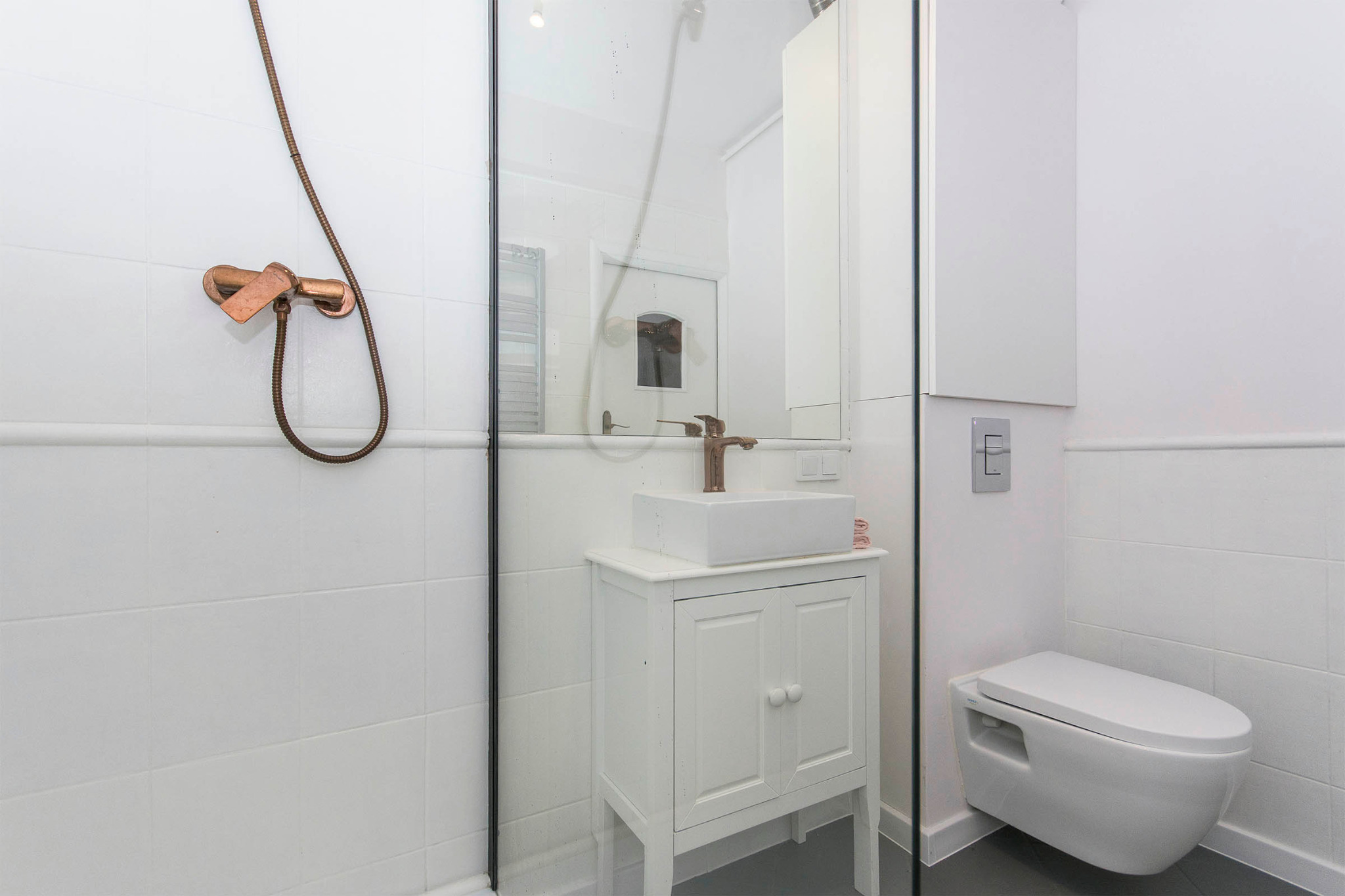 Nowoczesna łazienka z białą ceramiką, kabiną prysznicową ze szklaną ścianką, umywalką na szafce i armaturą w kolorze miedzi.