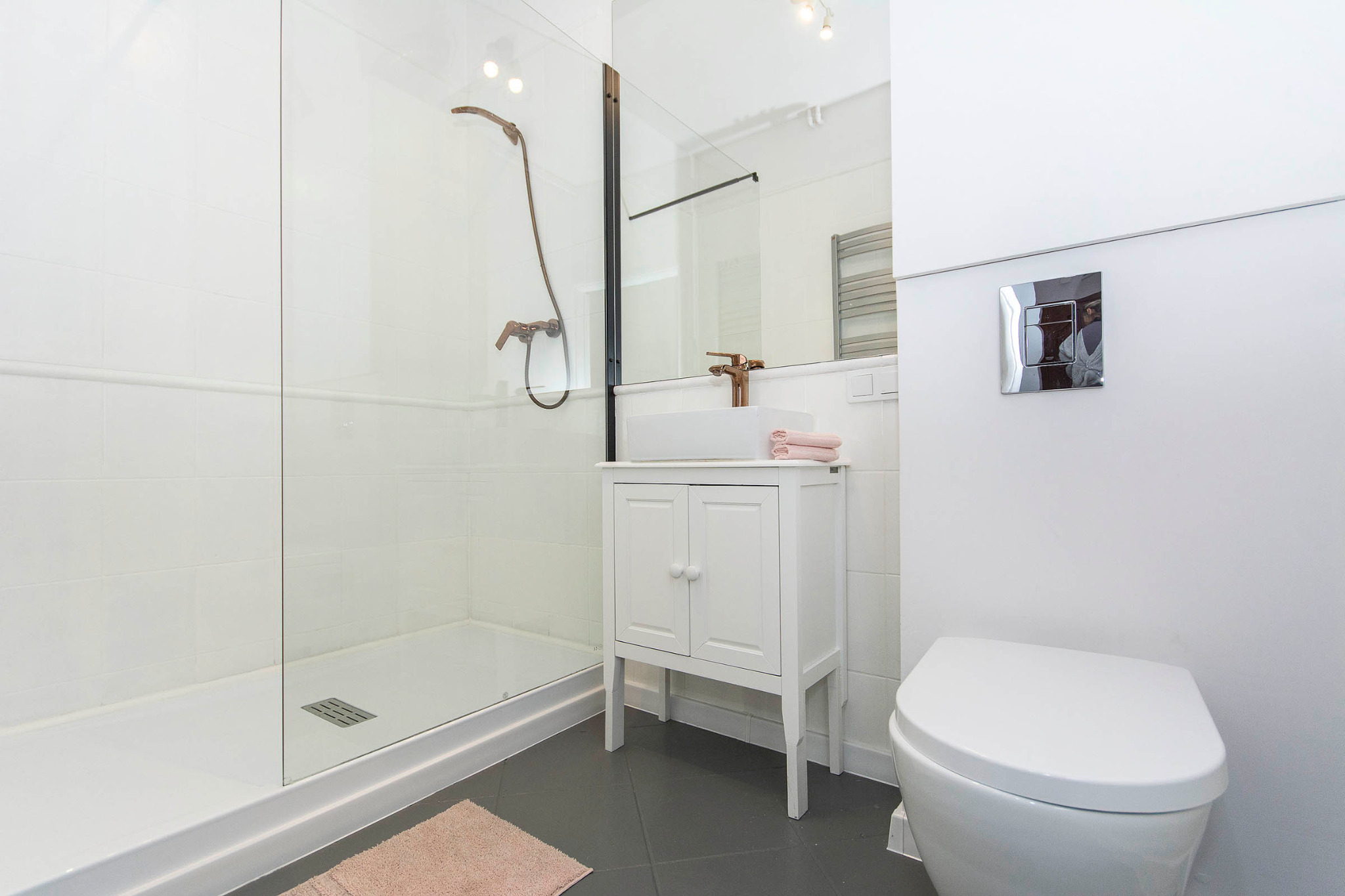 Nowoczesna łazienka z kabiną prysznicową typu walk-in, białą szafką z umywalką nablatową i armaturą w kolorze miedzi, uzupełniona o minimalistyczną toaletę i różowy dywanik.