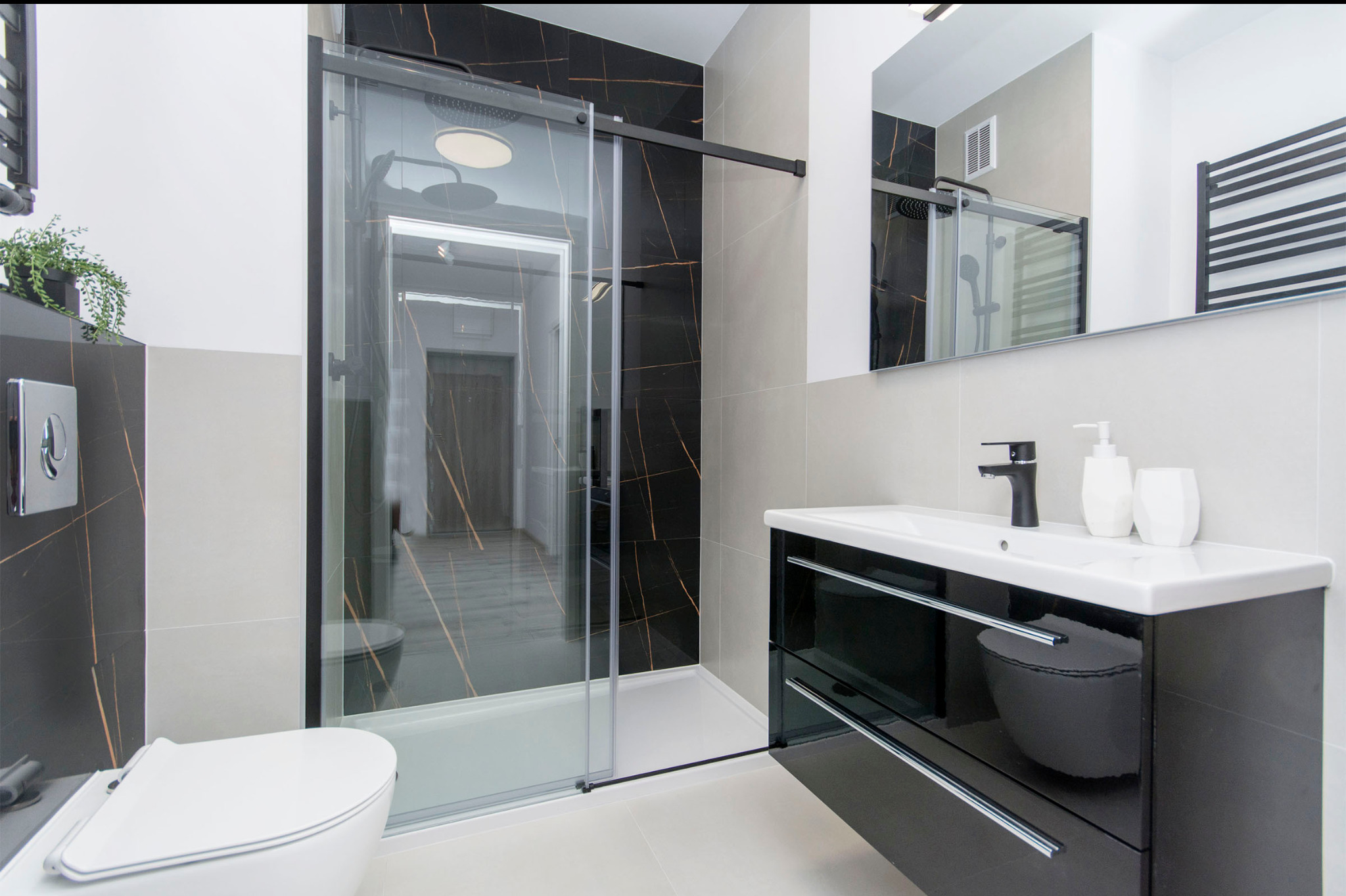 Nowoczesna łazienka z czarnymi marmurowymi akcentami, kabiną prysznicową typu walk-in z przesuwnymi drzwiami, umywalką nablatową na czarnej szafce z chromowanymi uchwytami i wiszącą toaletą.